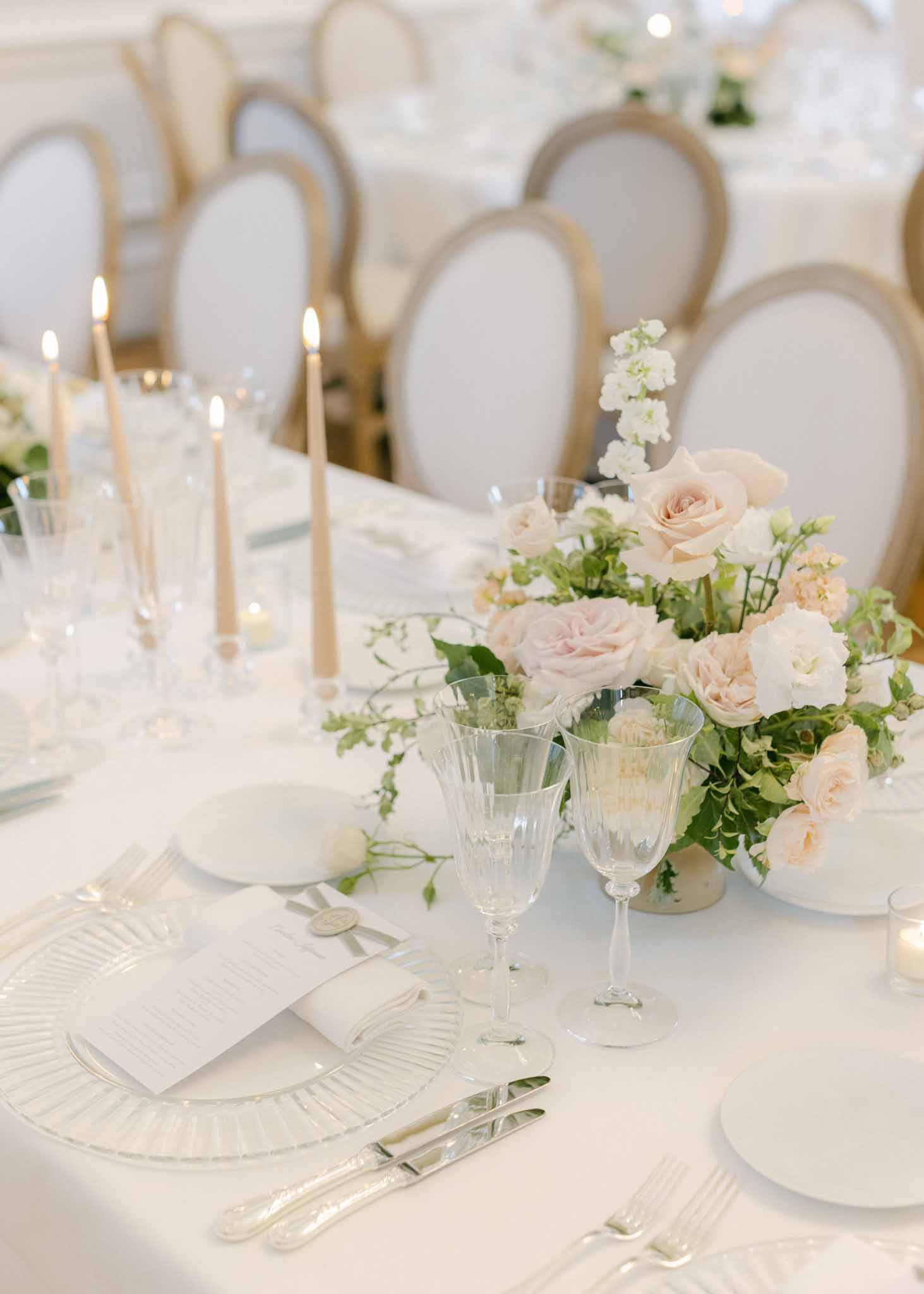 Reception table with blush and cream rose centerpiece in glass vase, white charger plates and cream taper candles