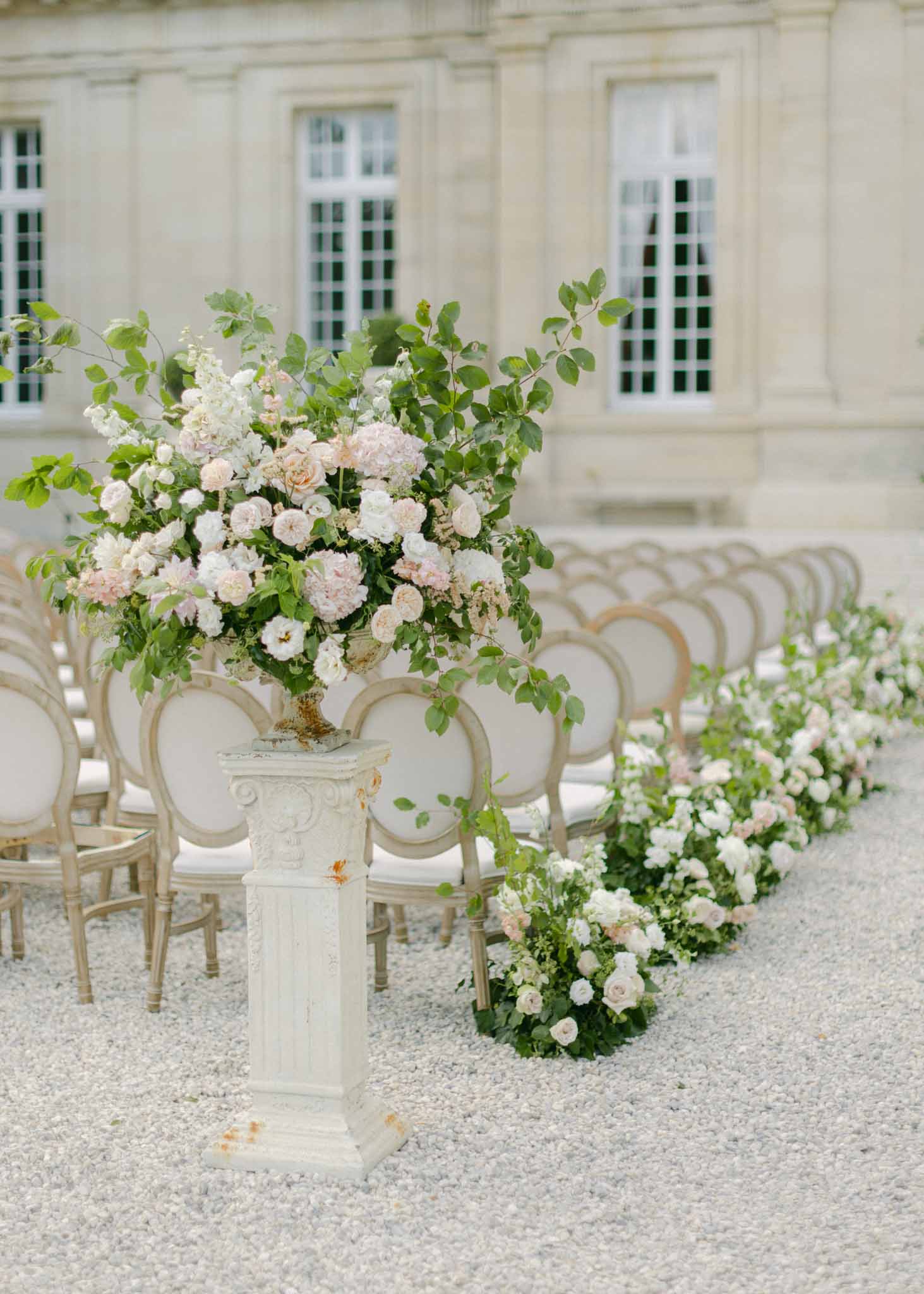 Outdoor ceremony aisle with ivory pedestals of blush roses and white hydrangeas beside classical stone building