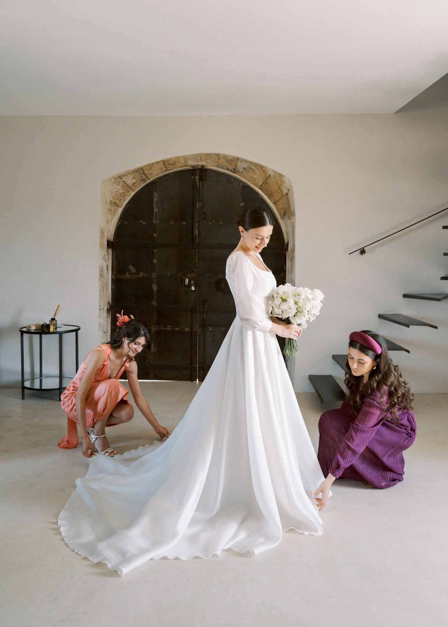 Bride in white puff-sleeve gown standing in arched doorway while two attendants arrange her cathedral train