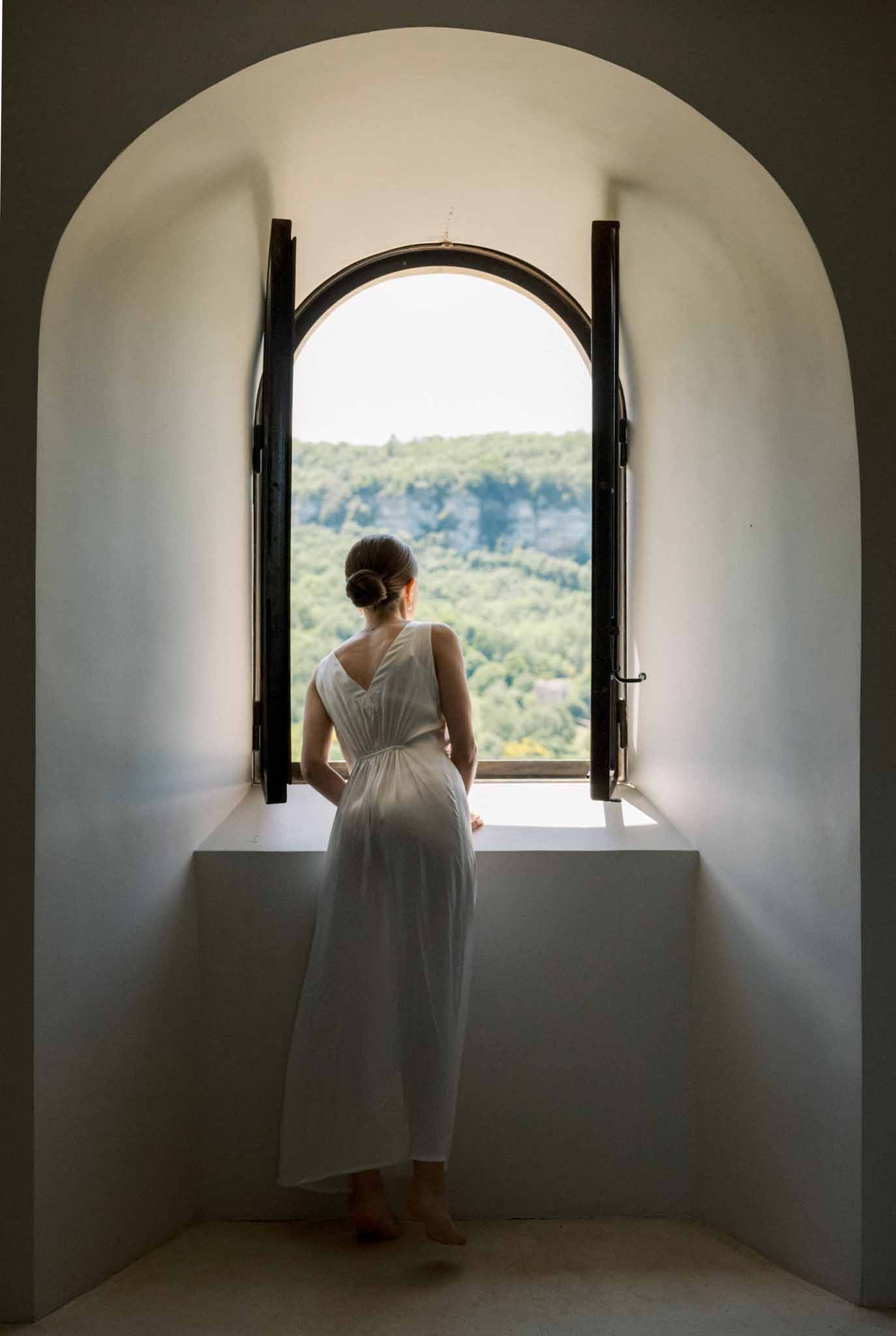 Bride in white sleeveless midi dress with low bun looking out through arched window in thick plastered chateau wall
