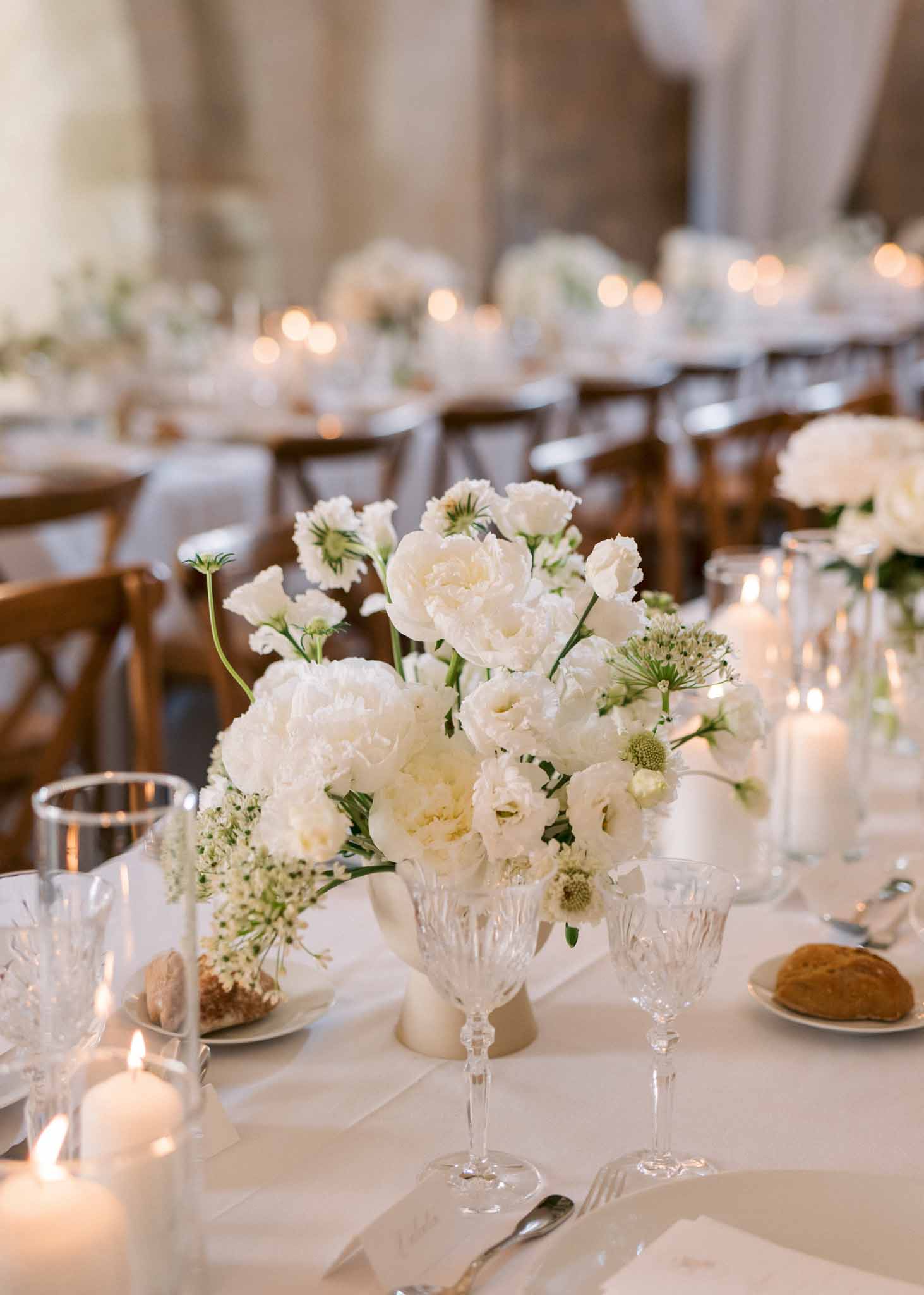 Reception table with white peony centerpiece in ceramic vase, crystal glasses, pillar candles, and cross-back chairs