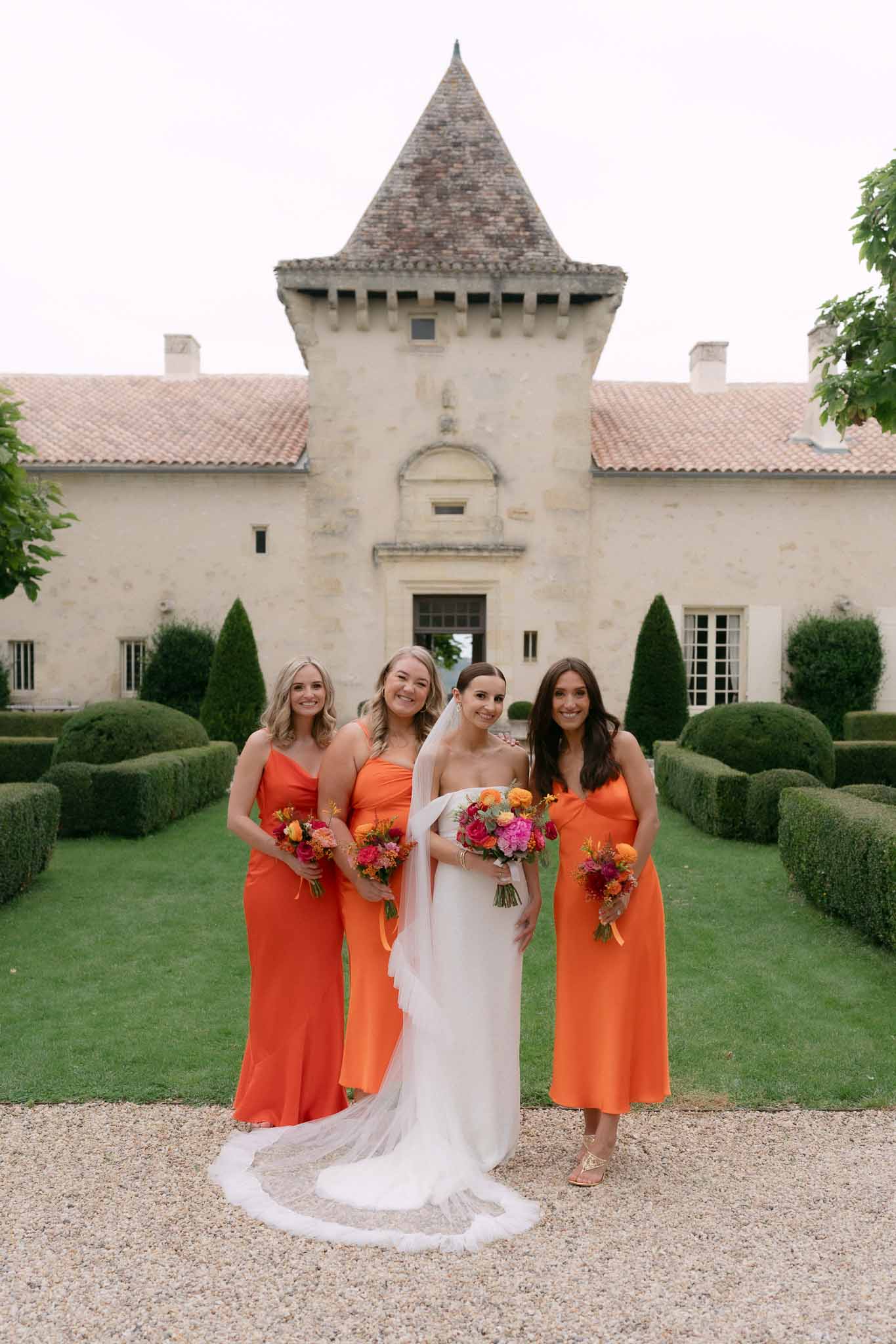 Bride in white strapless gown with three bridesmaids in tangerine orange satin dresses holding colorful bouquets at chateau