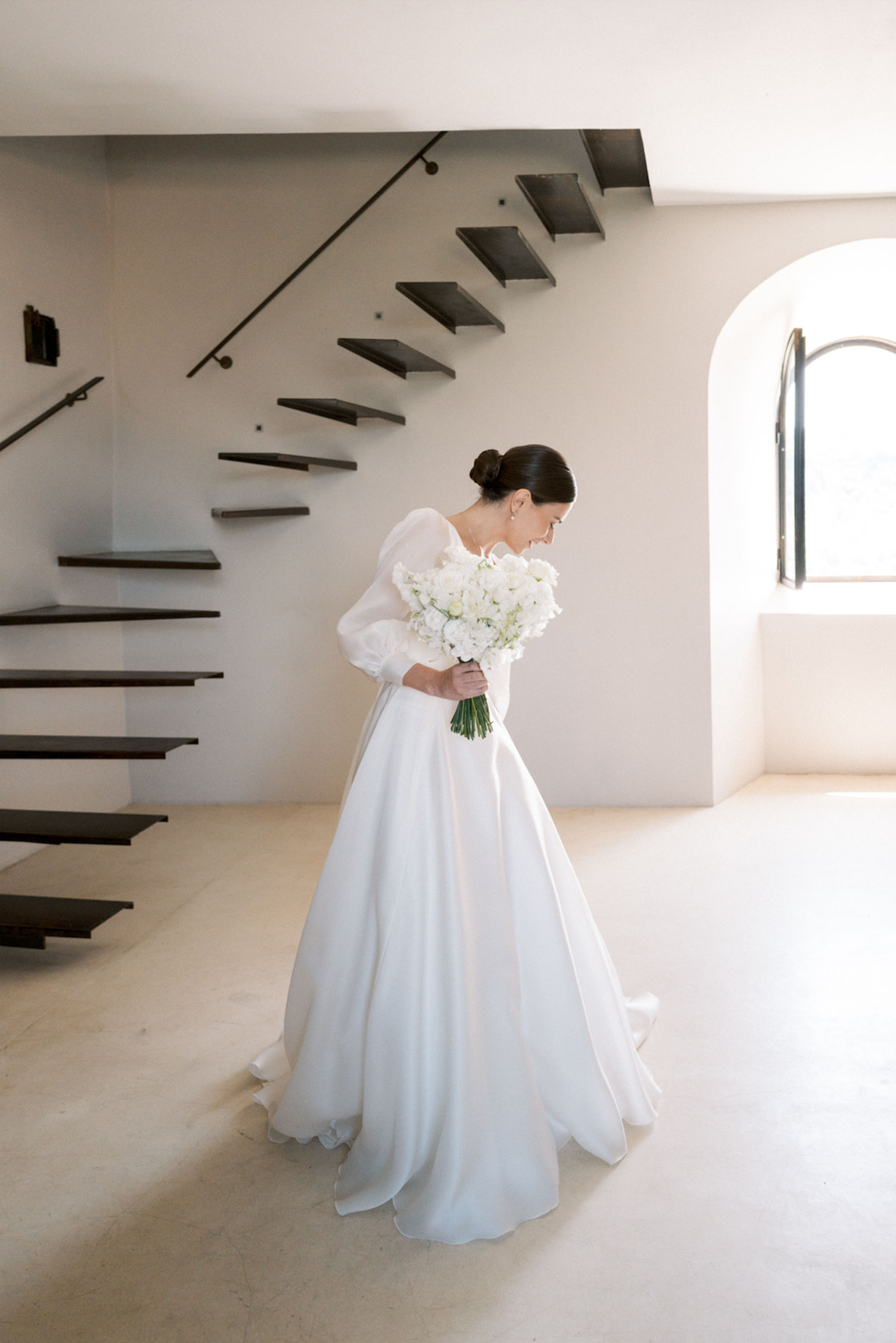 Bride in white puffed-sleeve ball gown holding all-white bouquet in minimalist interior with floating steel staircase