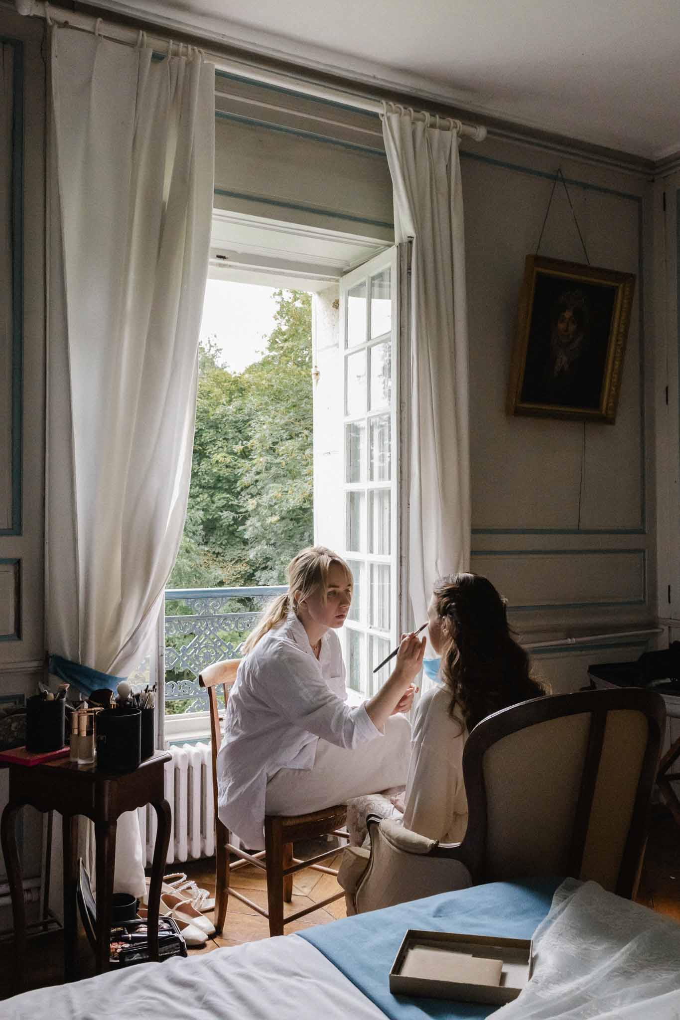 Bride having makeup applied in classical French bedroom during wedding preparation