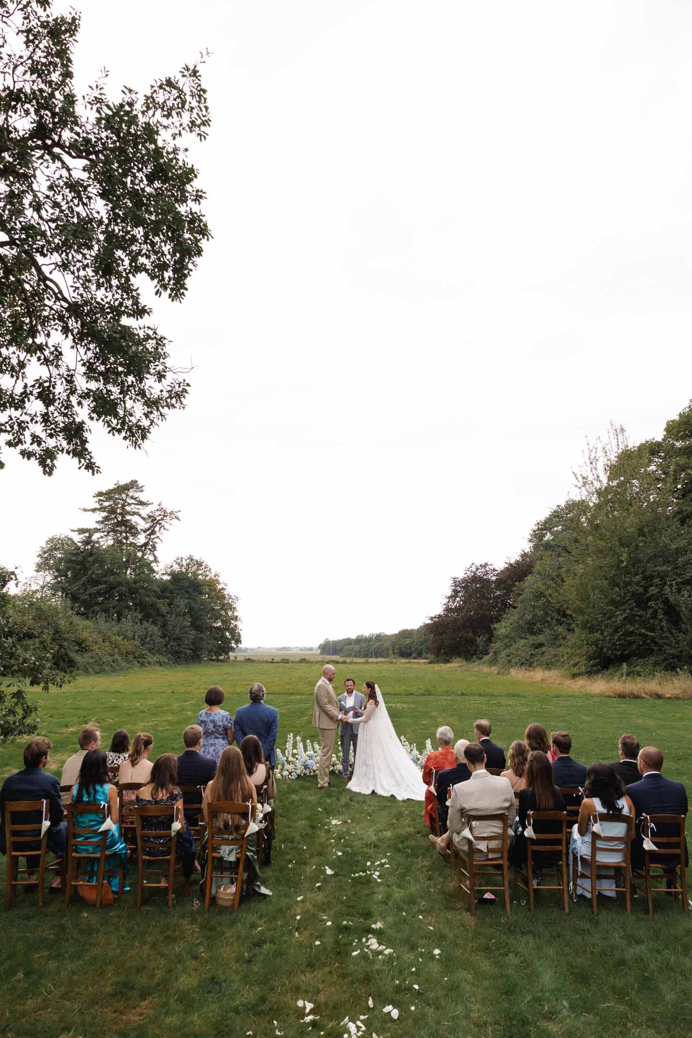 Outdoor wedding ceremony in open field with guests seated on wooden chairs facing couple and officiant