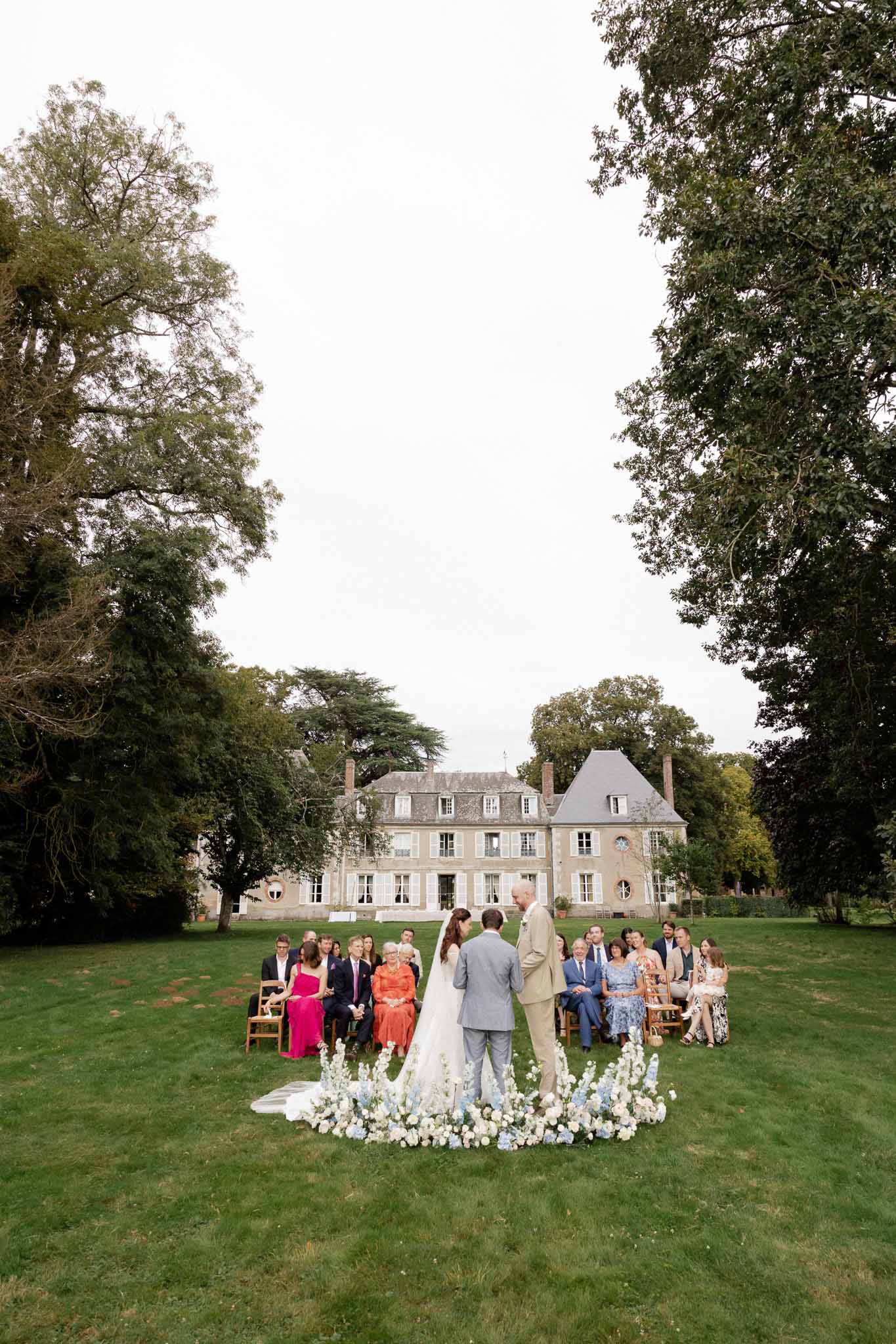Wedding ceremony with circular floral arch on chÃƒÂ¢teau lawn with guests in wooden chairs