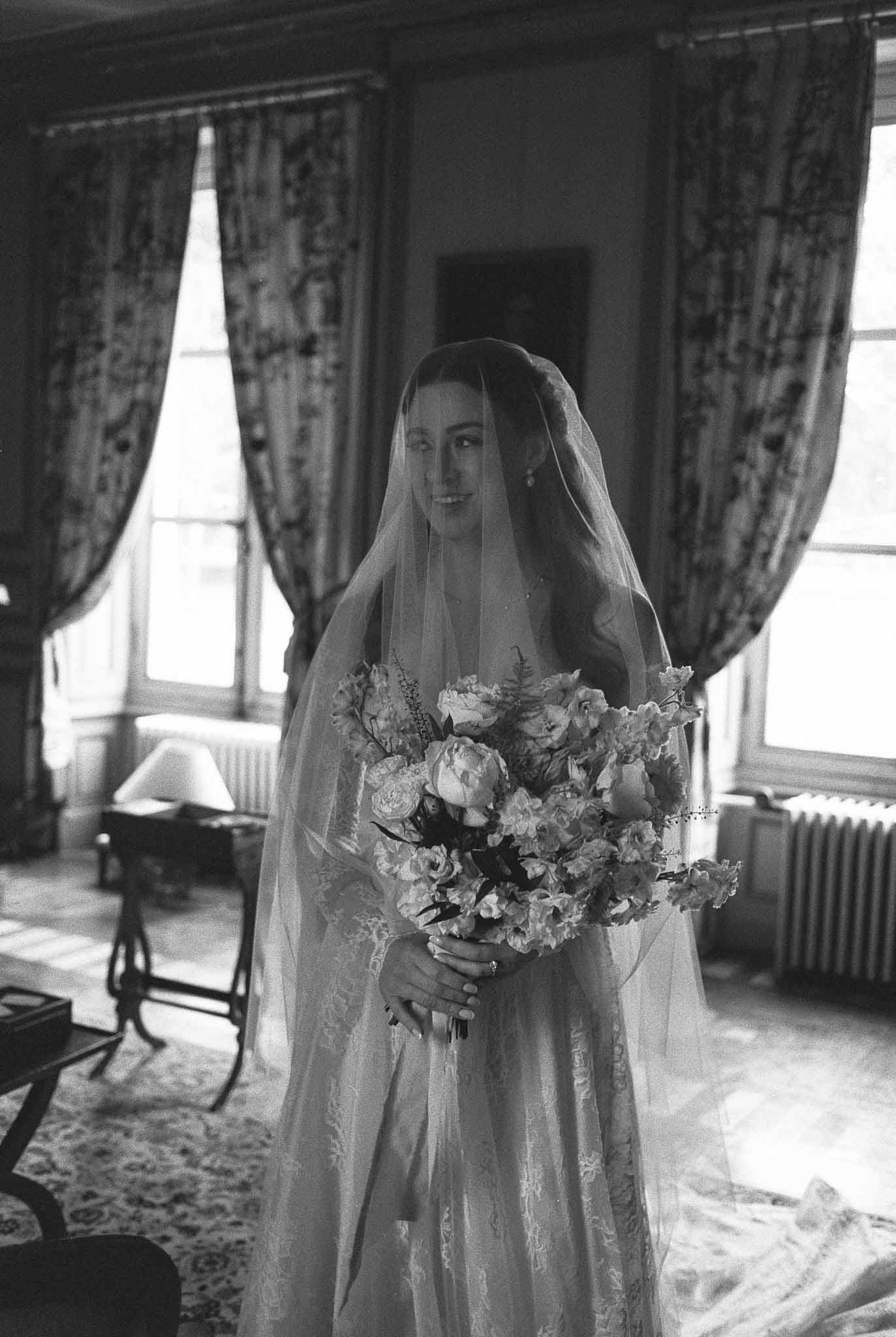 Bride in wedding dress and veil holding bouquet in formal indoor setting with tall windows