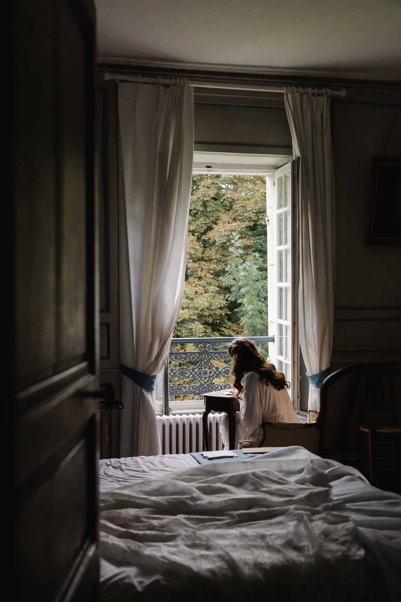 Bride in ivory dress sitting on bed gazing through French doors at European venue during getting ready
