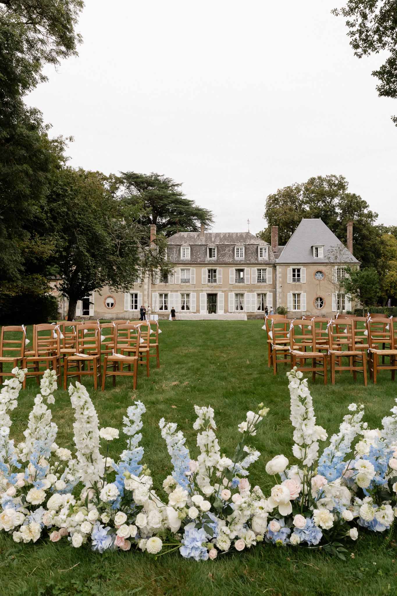 Outdoor wedding ceremony setup on lawn of French chÃƒÂ¢teau with floral aisle borders and cross-back chairs
