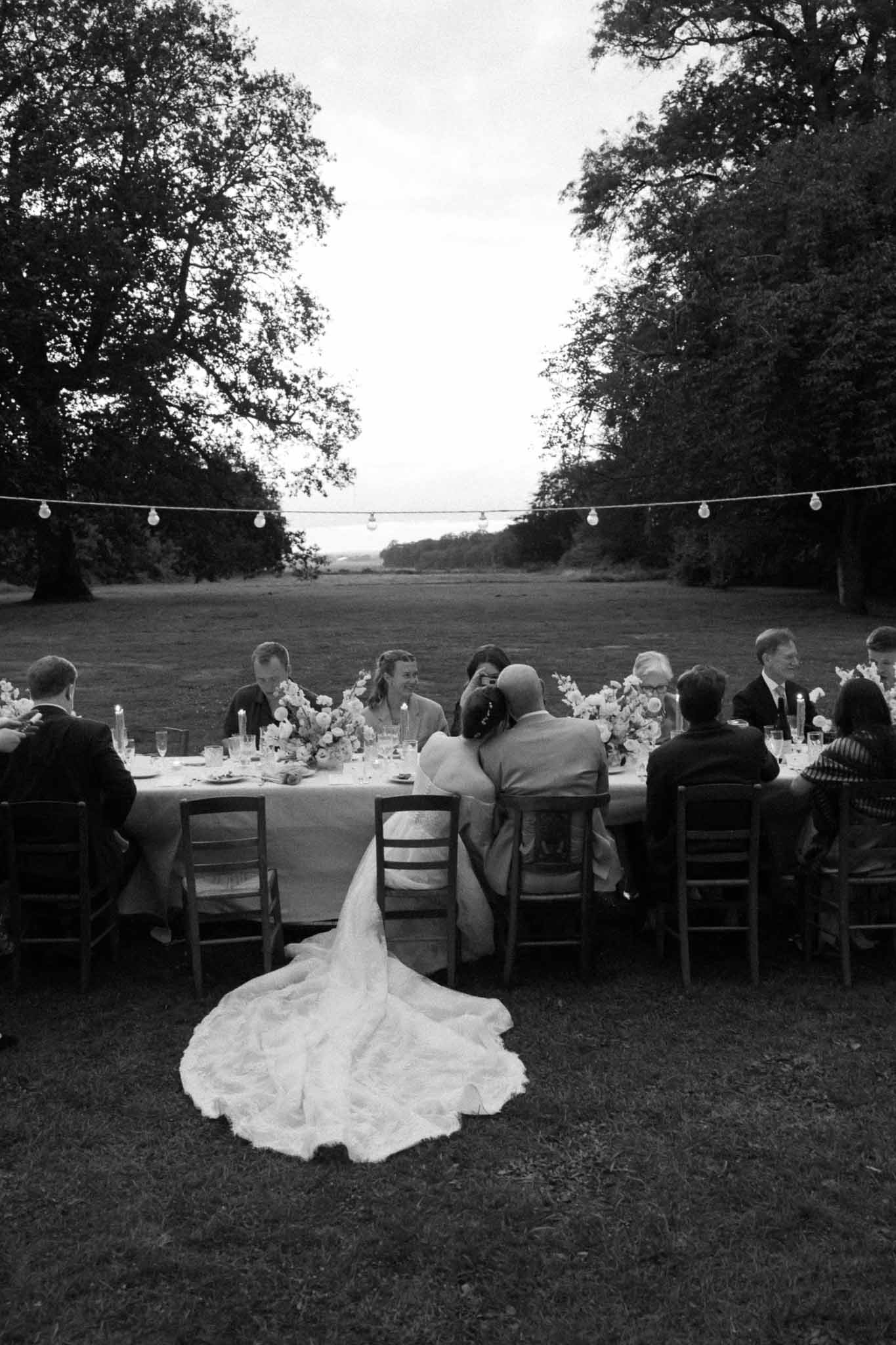 Outdoor reception dinner with bride and guests at long table under trees with string lights