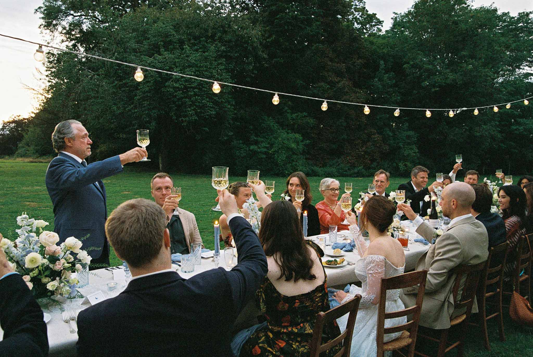 Wedding reception speeches with guests raising glasses at outdoor dinner table with string lights and floral centerpieces