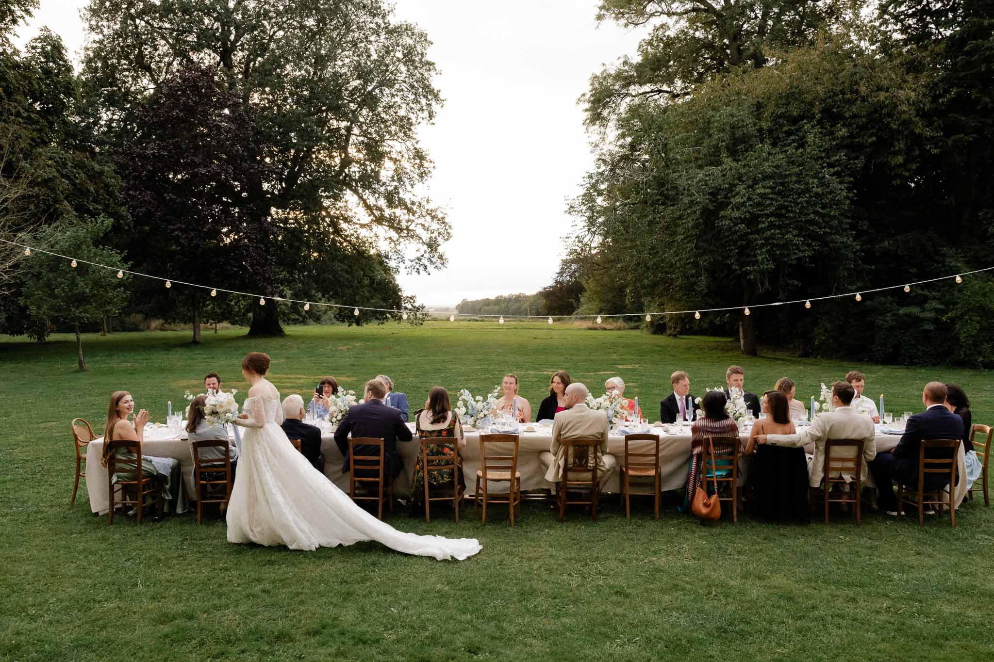 Bride and guests at long outdoor reception table in garden setting with string lights