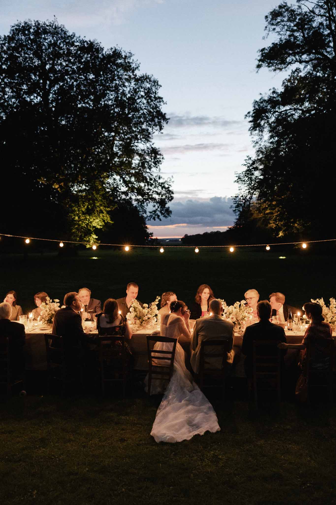 Outdoor reception dinner with guests seated at long banquet table under string lights at dusk in countryside setting