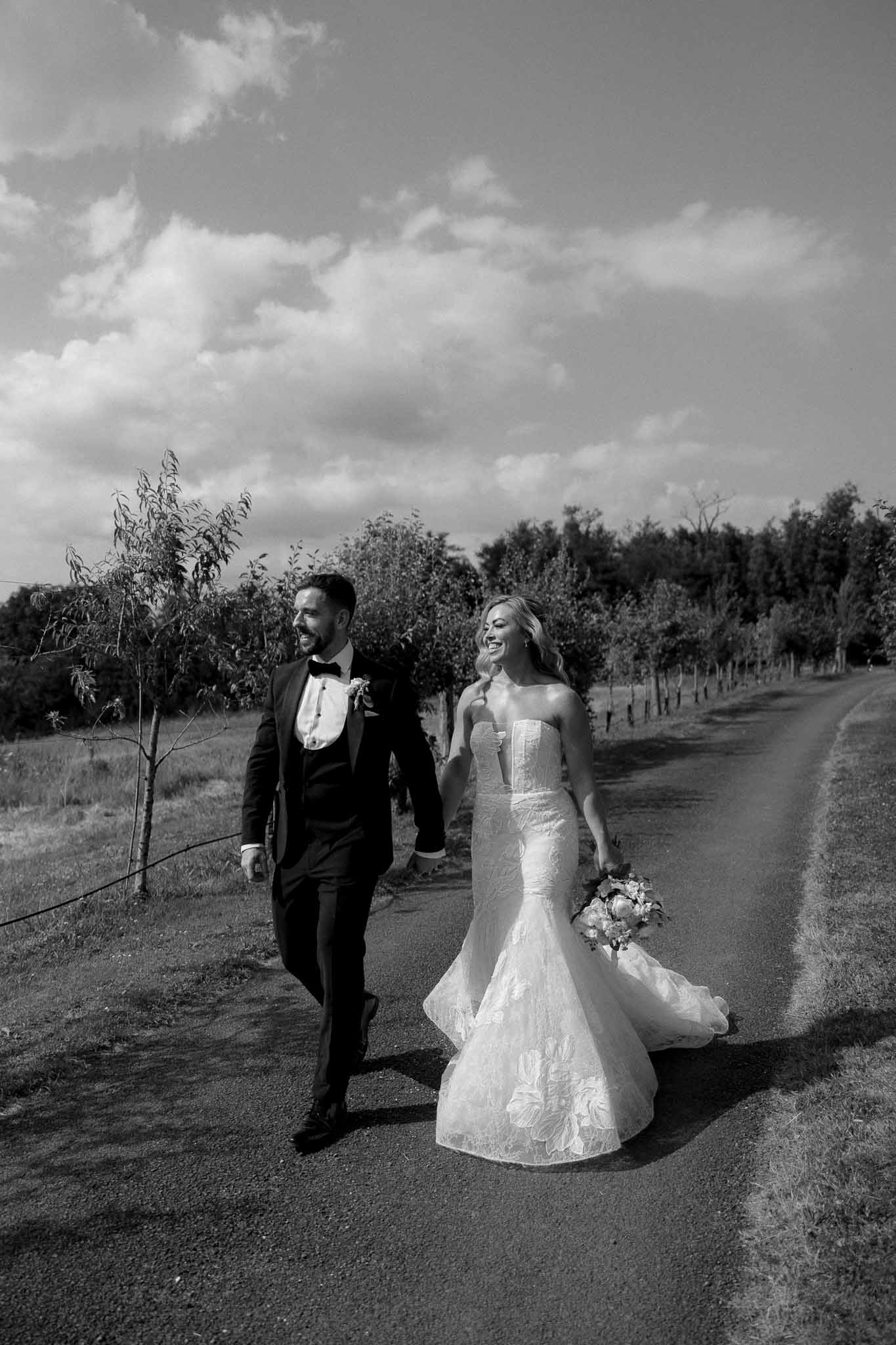 Bride and groom walking together down rural vineyard driveway in black and white portrait