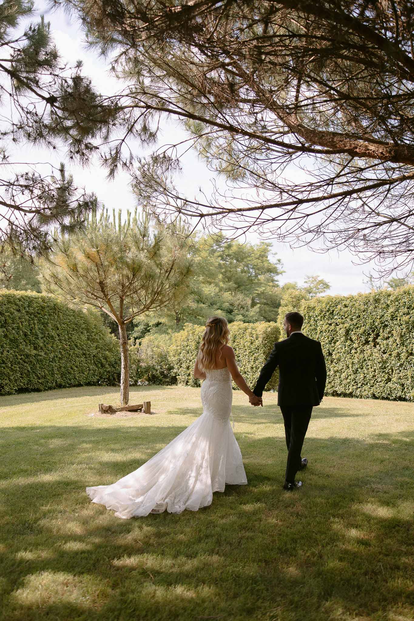 Bride and groom walking hand-in-hand through formal garden with manicured hedges and ornamental tree