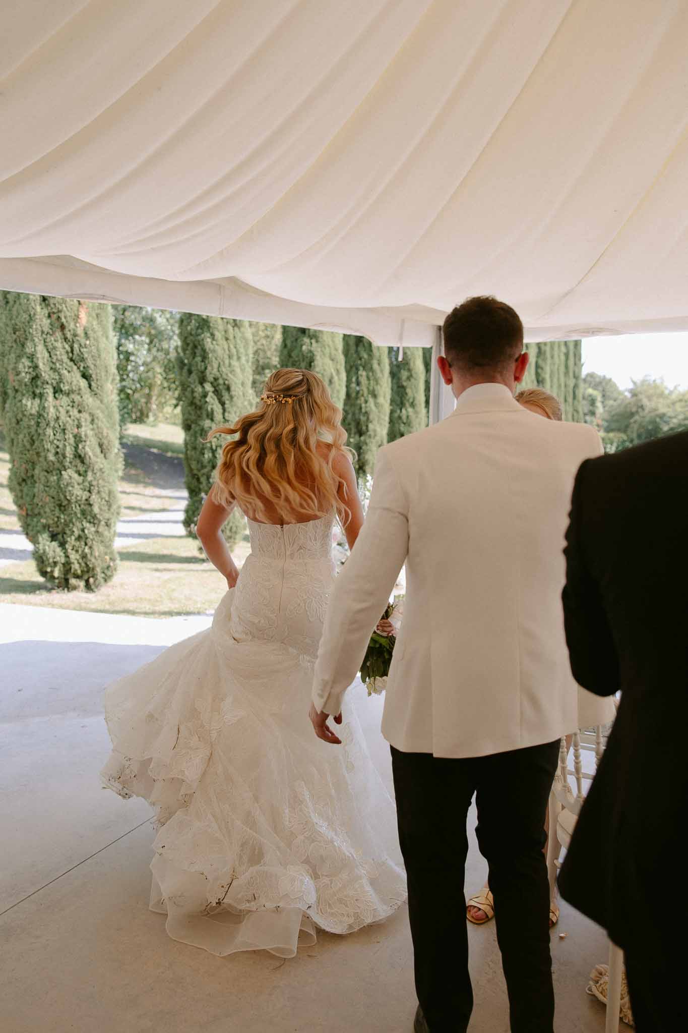 Bride and groom walking away under white marquee, bride in strapless lace mermaid gown with gold hair accessory