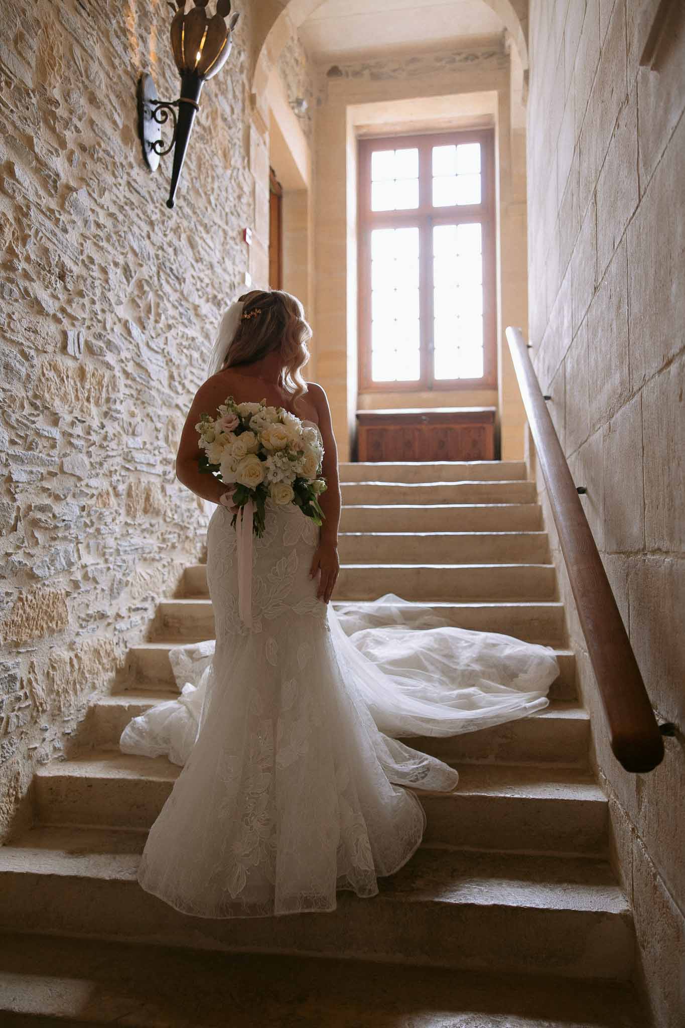 Bride in ivory lace wedding dress ascending stone staircase in historic castle interior
