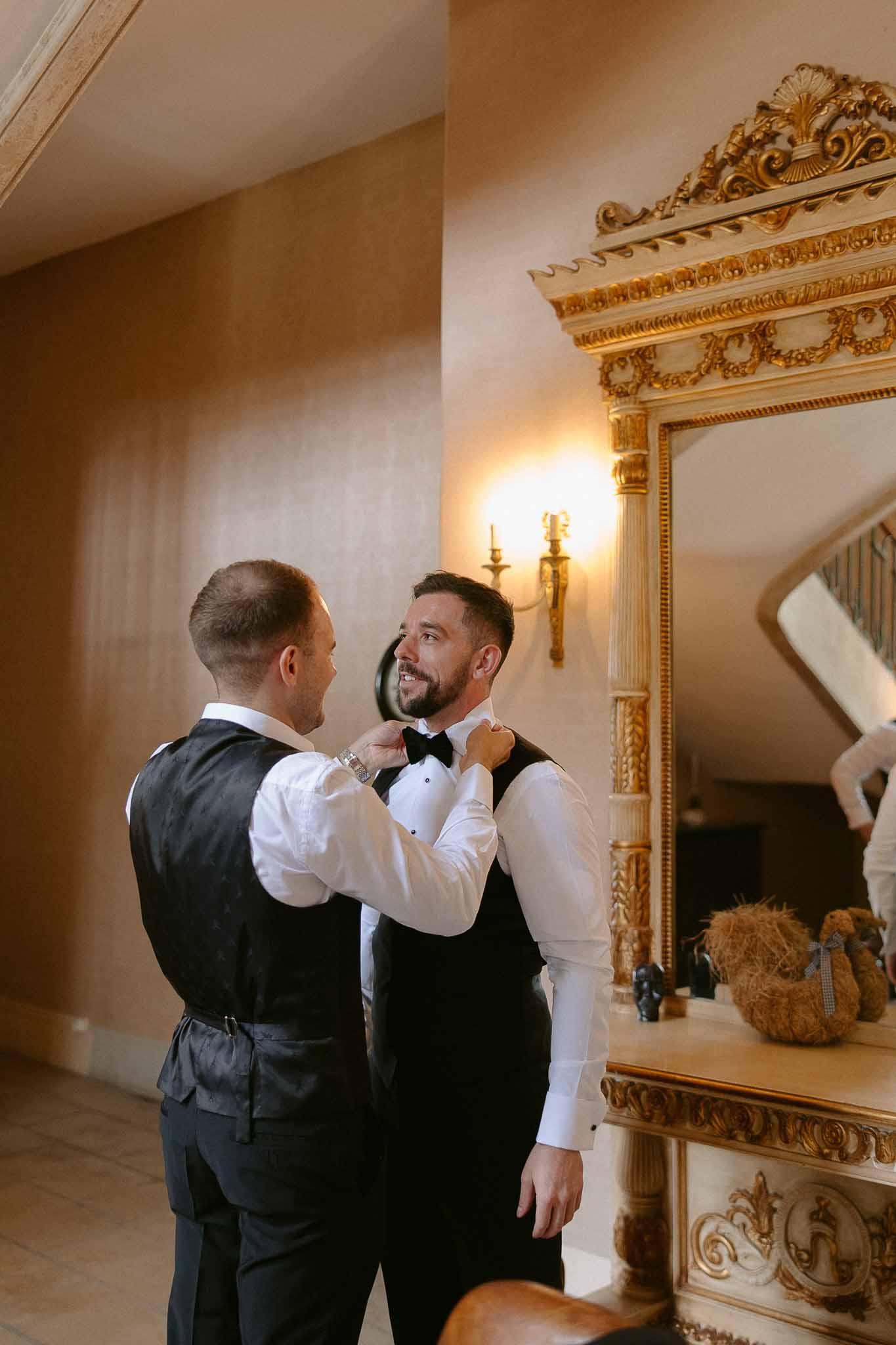 Groom being helped with bow tie by groomsman in elegant classical interior with ornate gold molding