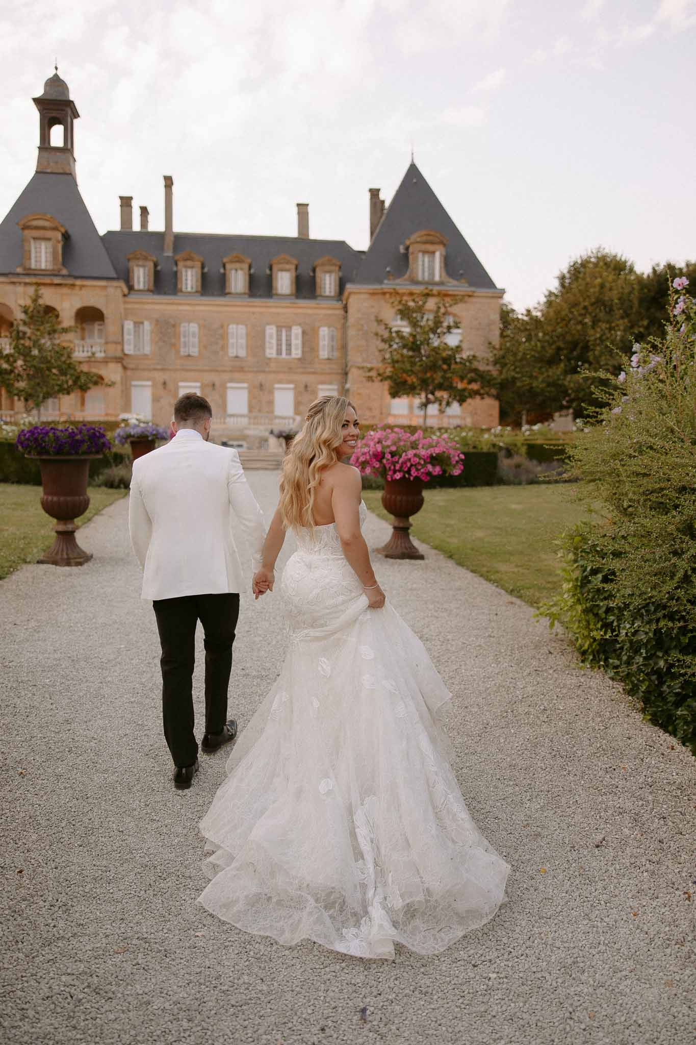 Bride and groom walking hand-in-hand toward grand brick chÃƒÂ¢teau with formal gardens