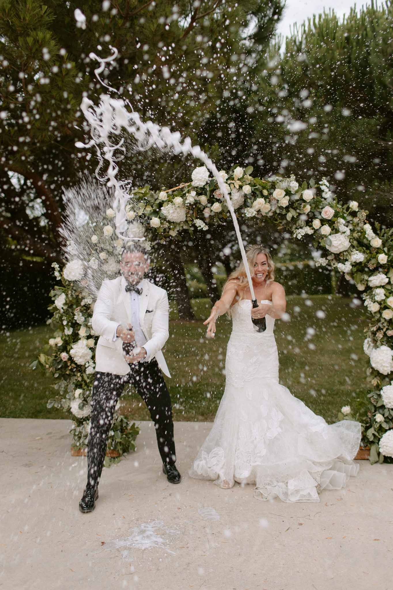 Bride and groom spraying champagne bottles on a terrace in front of a circular rose-covered floral arch