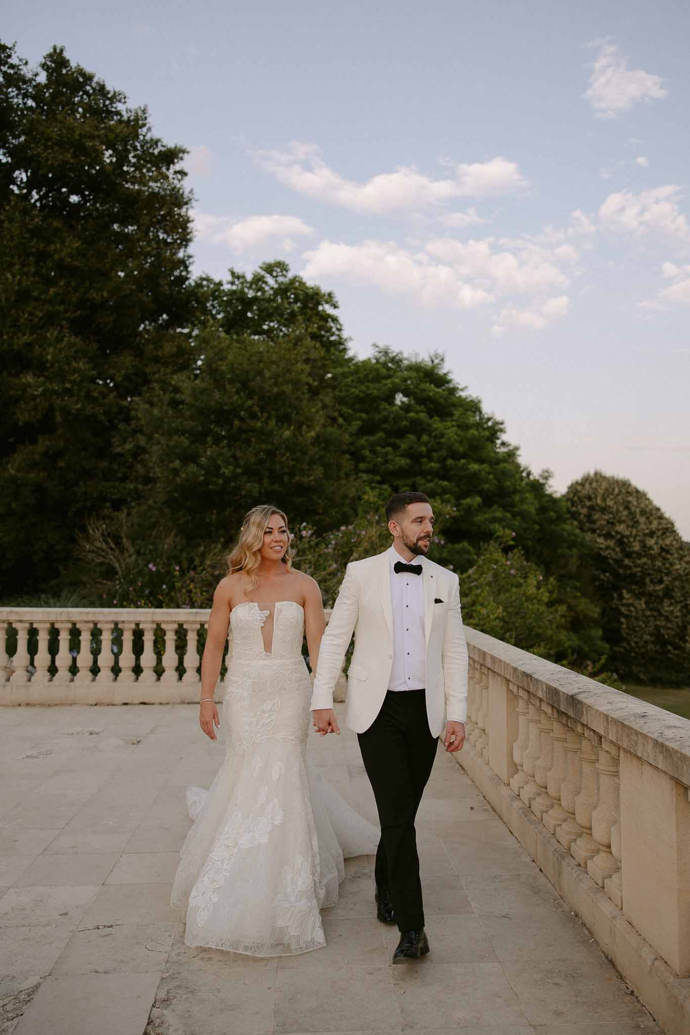 Bride and groom walking hand-in-hand on stone terrace overlooking manicured gardens