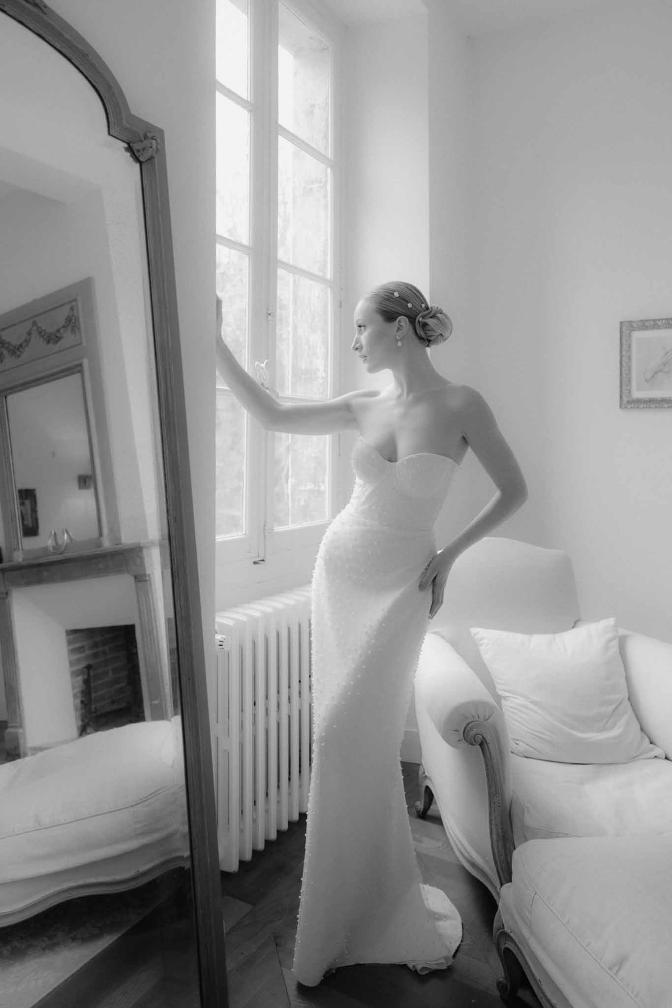 Bride in wedding gown looking out window in elegant bedroom during getting ready