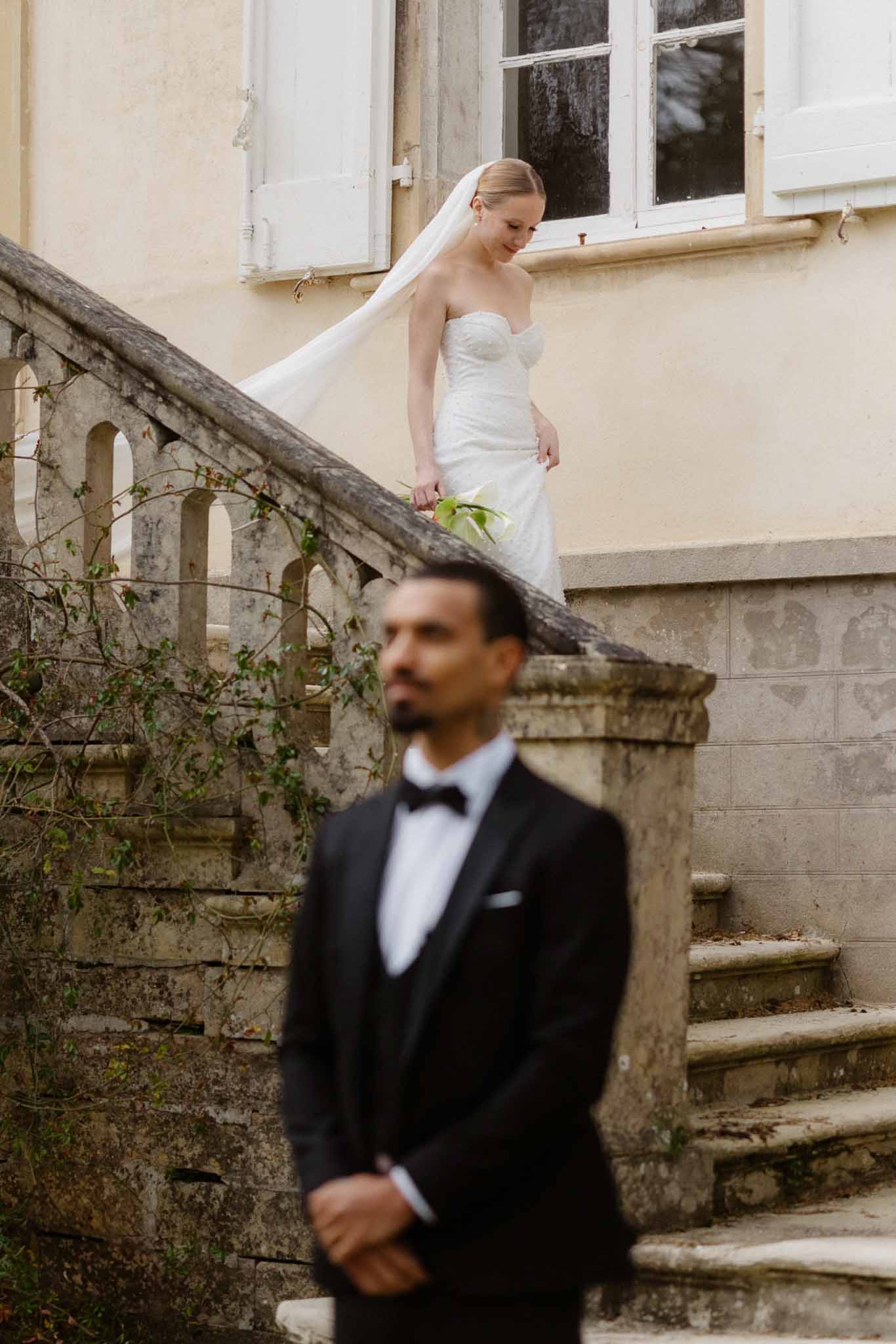 Bride in ivory dress descends stone staircase as groom waits below at European-style courtyard wedding venue