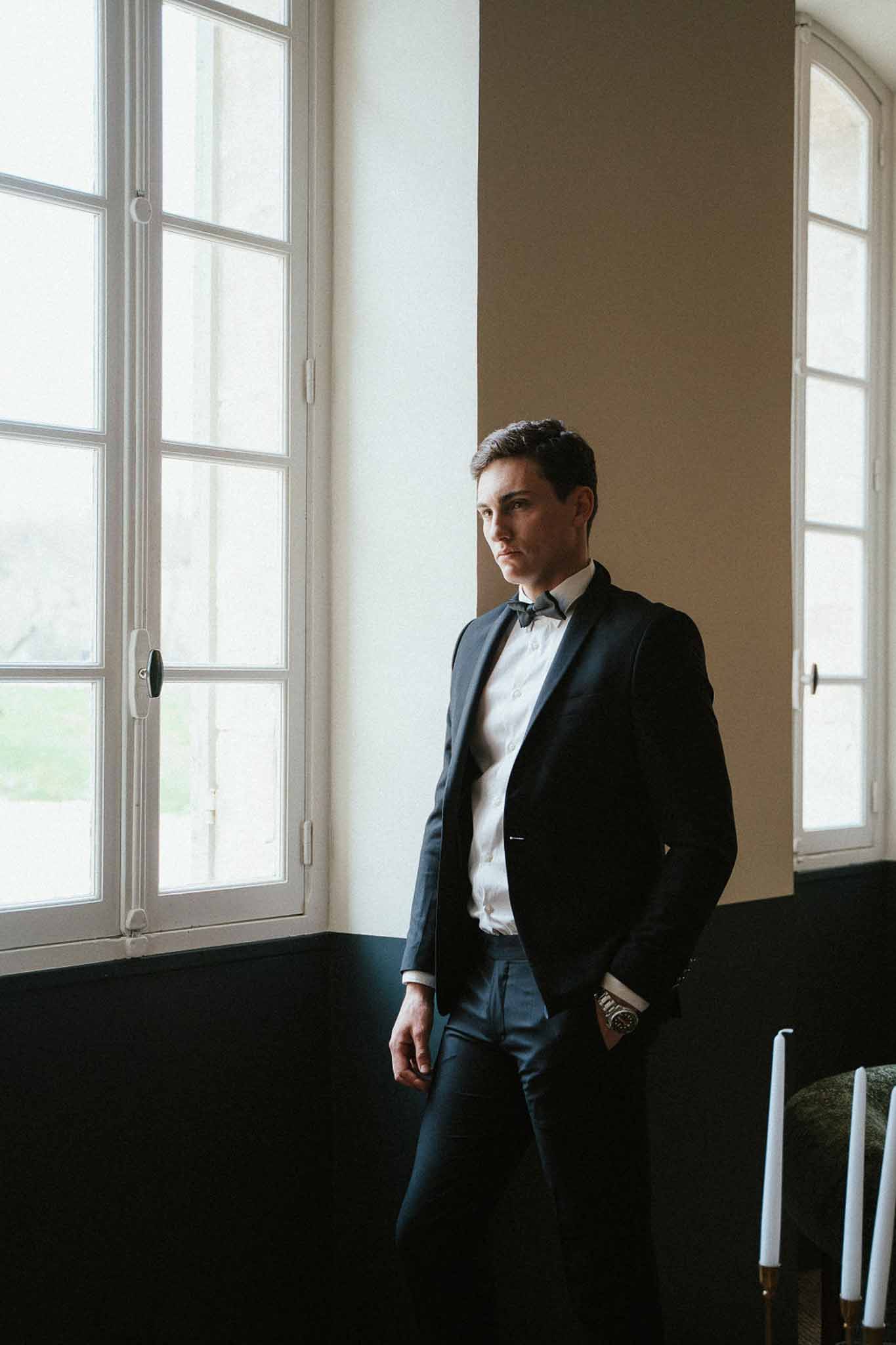 Groom in navy suit and bow tie standing between French windows with directional natural light and gold candles