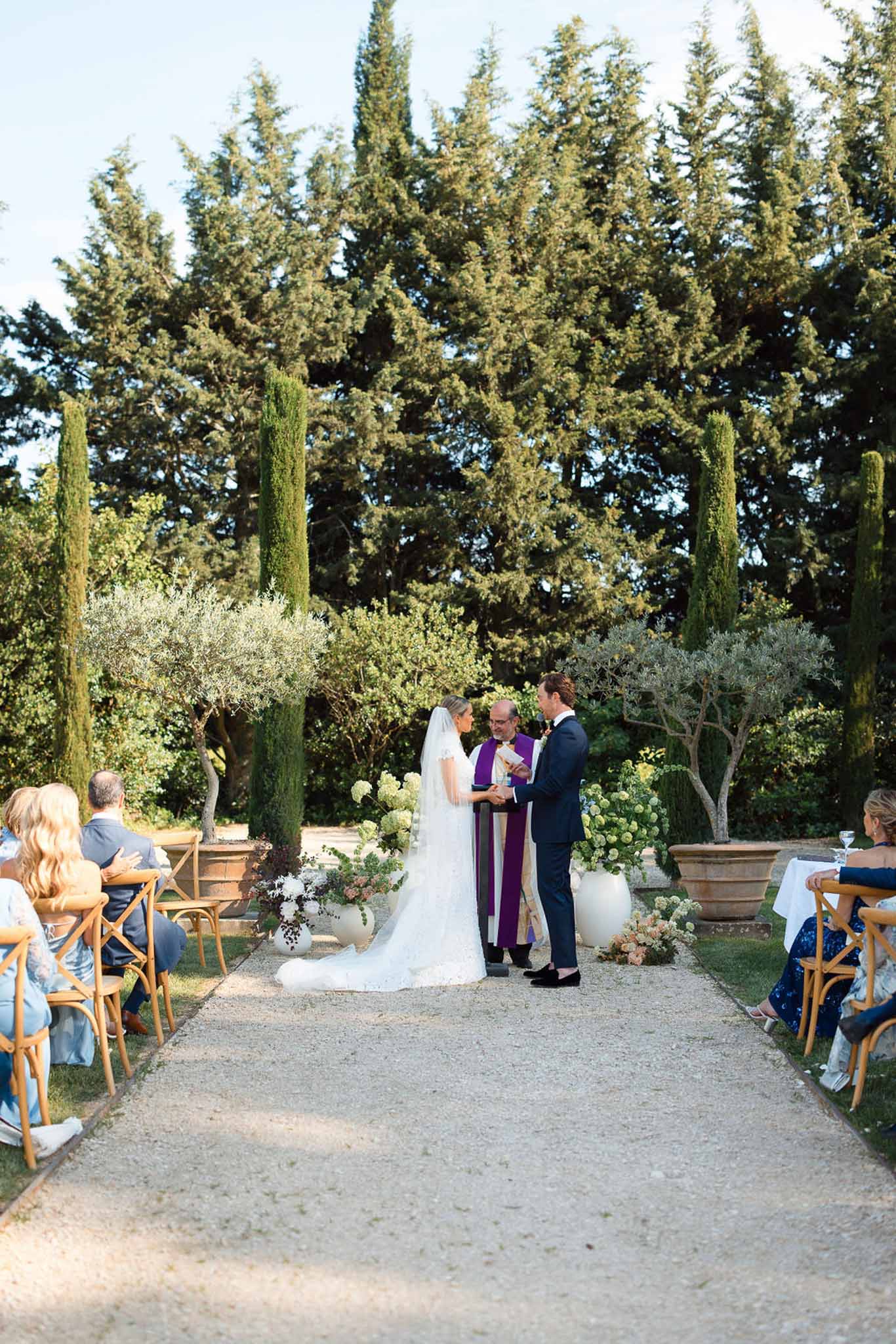 Wedding ceremony in Mediterranean garden with guests seated on natural wood chairs among cypress trees