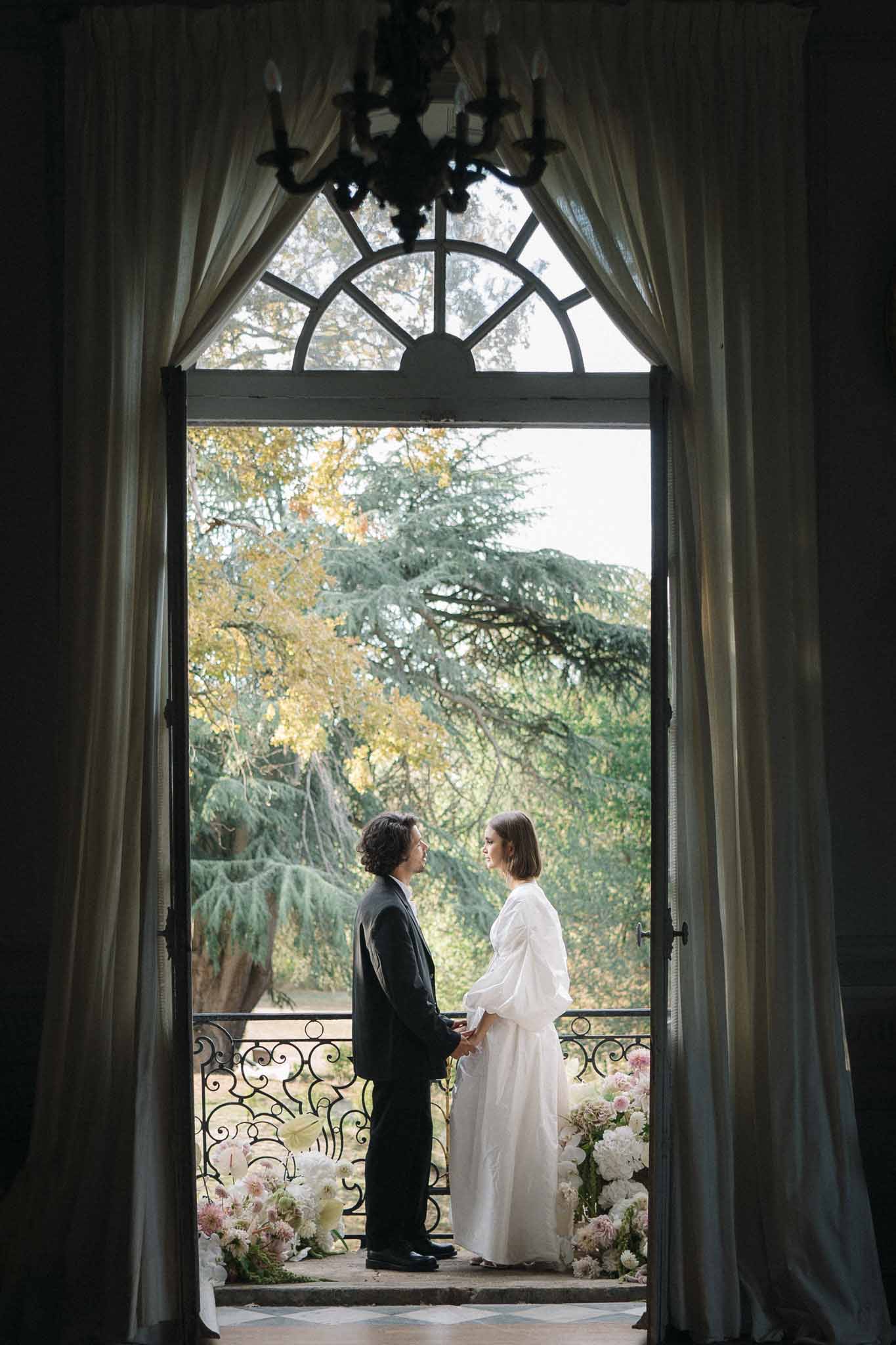 Couple holding hands at balcony doorway framed by draped curtains with blush dahlia and peony floor arrangements