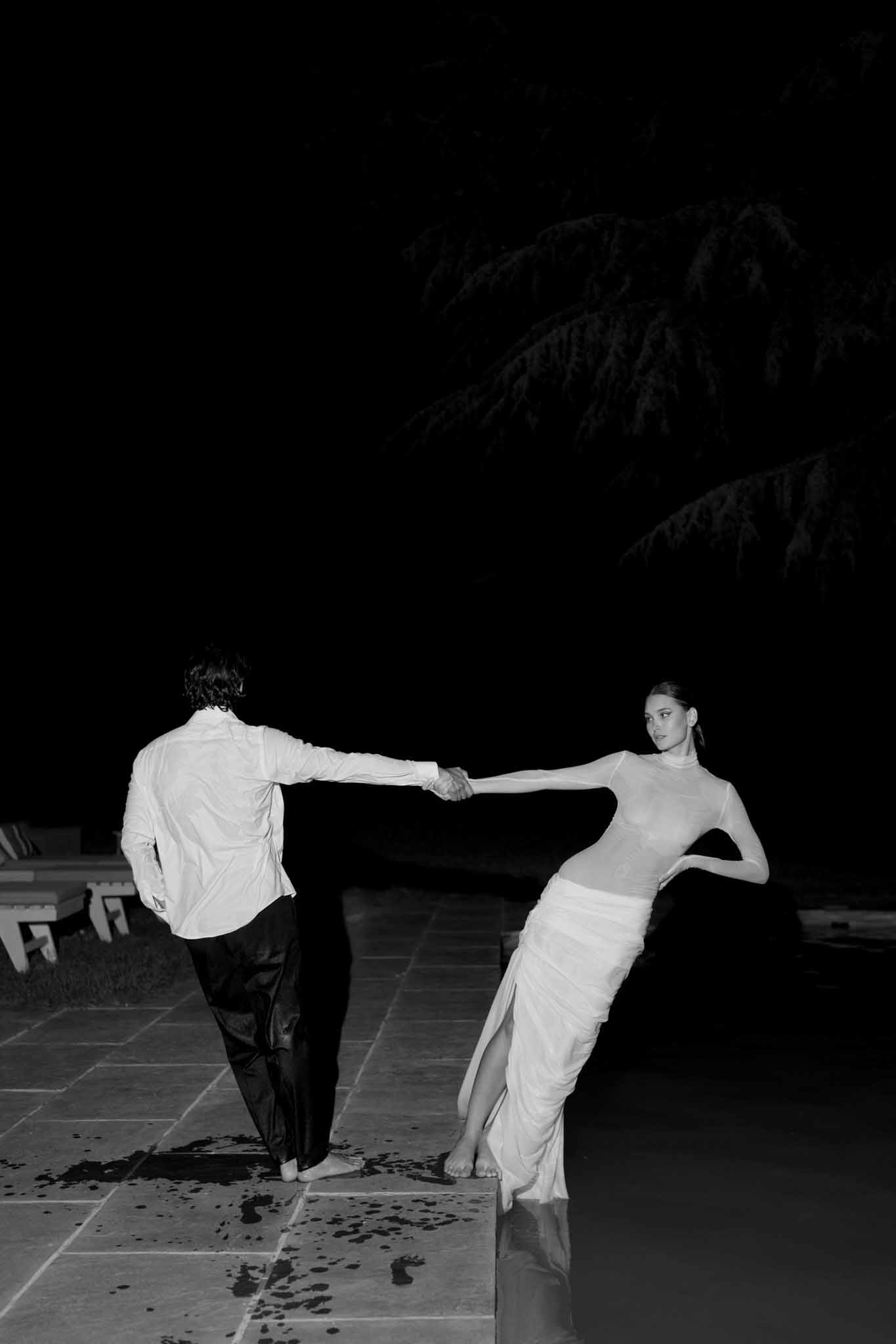 Black and white nighttime portrait of barefoot couple balancing at pool edge in dramatic contrast