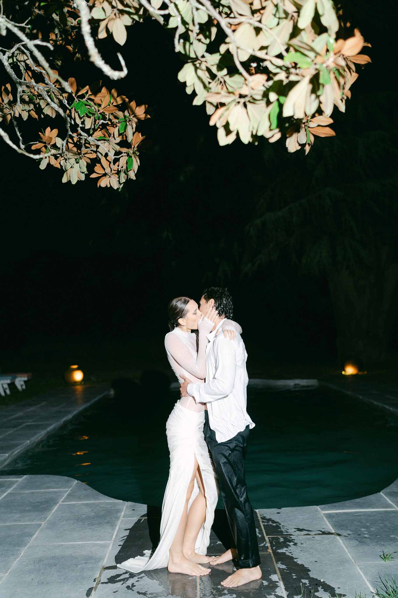Bride and groom kiss barefoot beside outdoor pool at night with amber ground lights reflecting on water