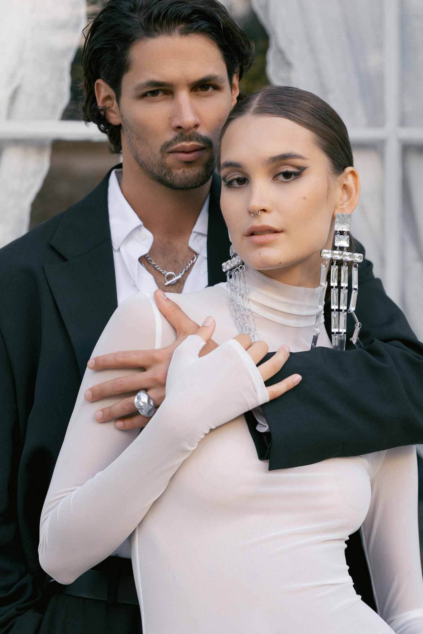 Bride in ivory turtleneck dress with crystal earrings embraced by groom in black blazer outdoors