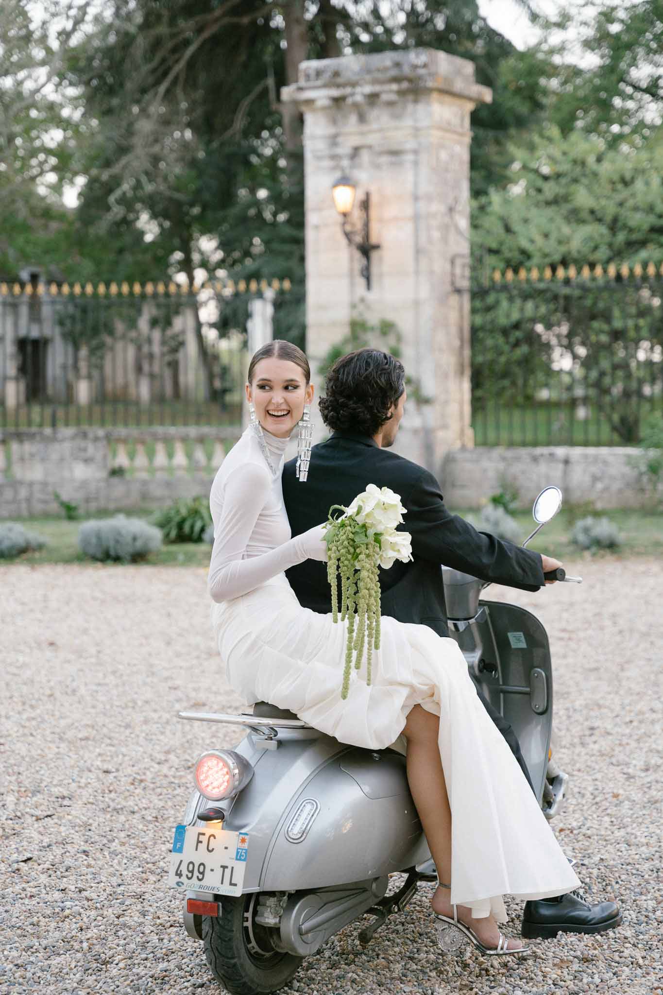 Bride and groom riding silver Vespa on chateau driveway bride looking back smiling in minimalist white gown