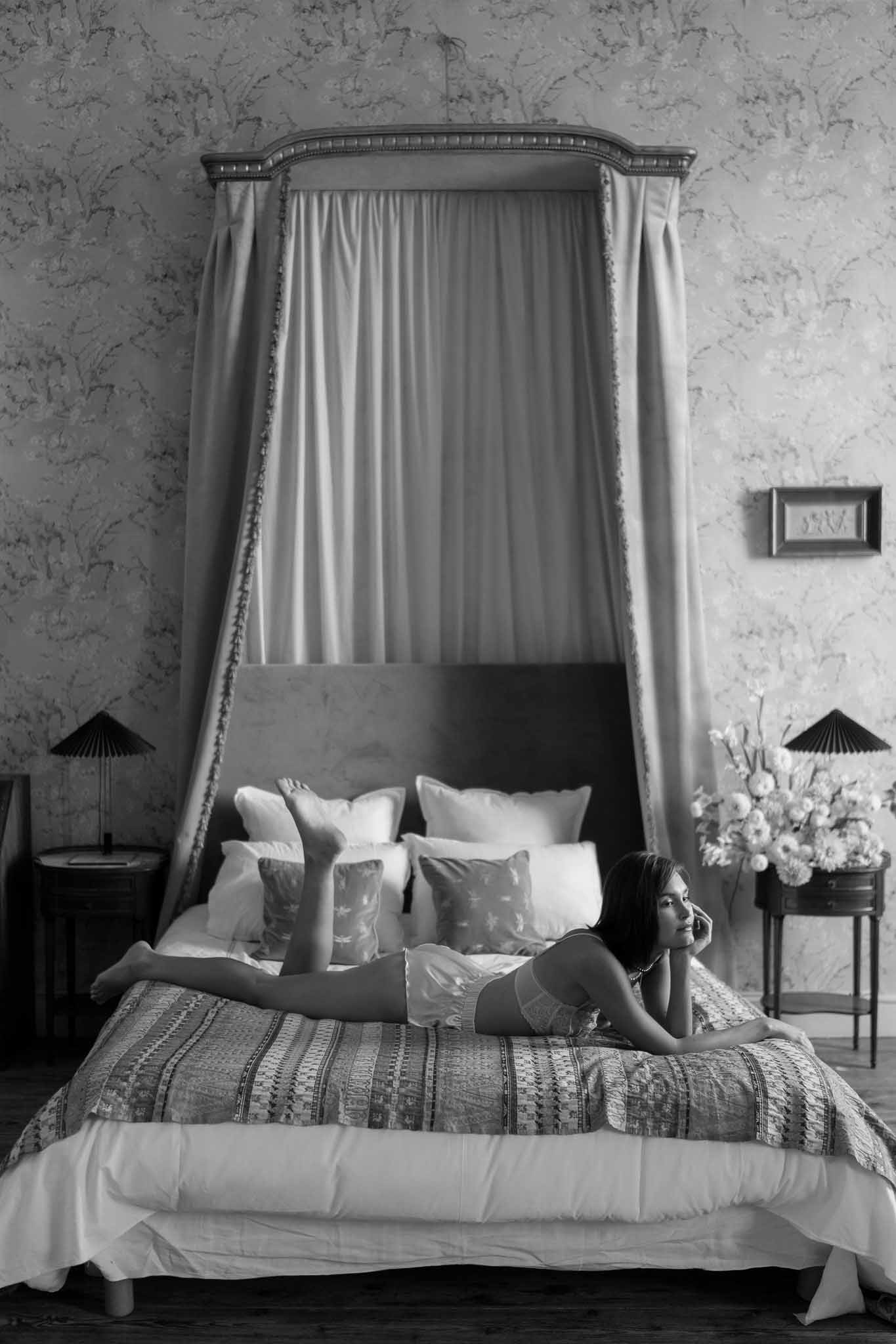 Black and white portrait of bride in lace lingerie lying on canopied bed in ornately decorated chateau bedroom