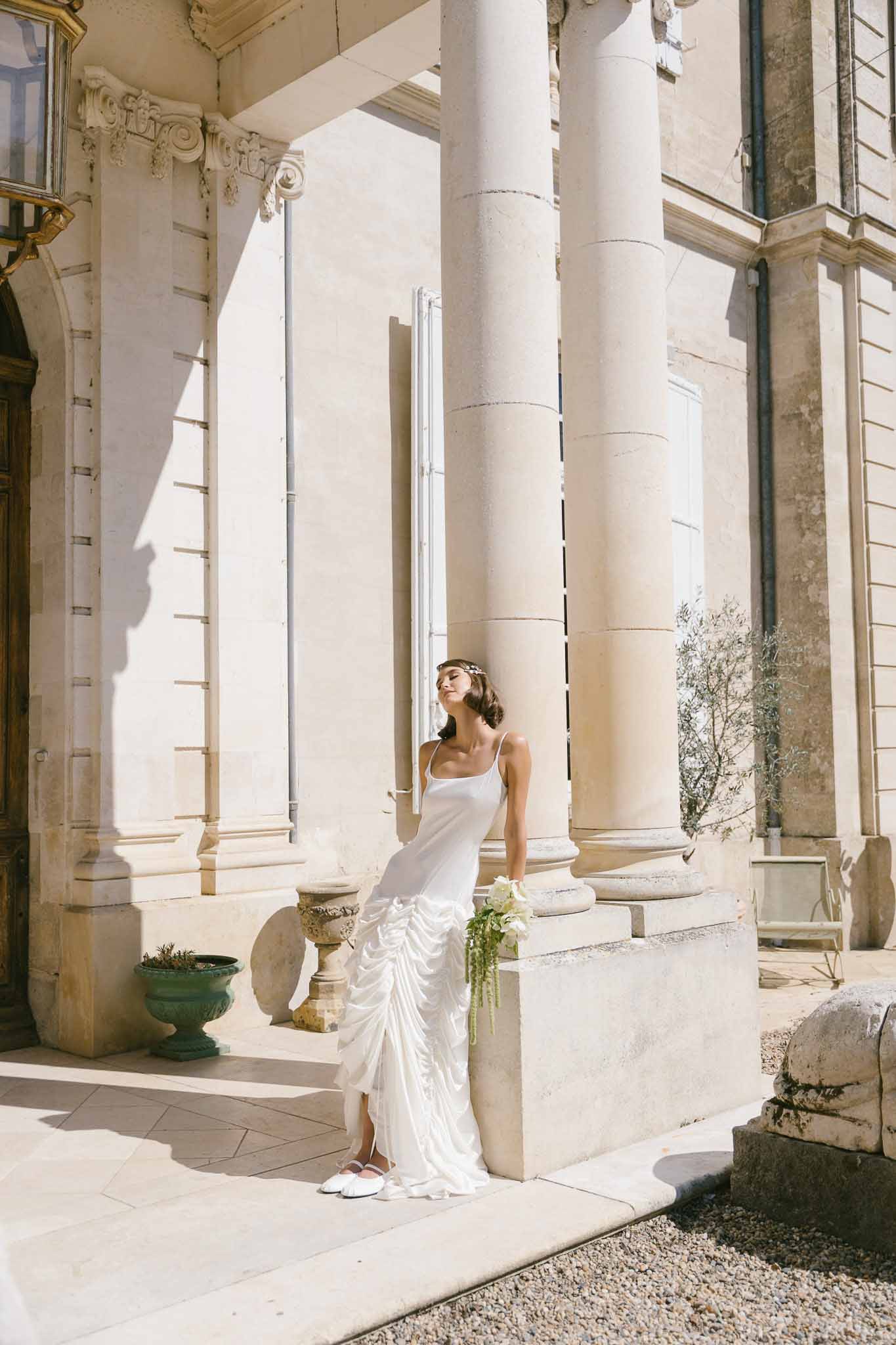 Bride in white ruffled slip gown with floral headband and trailing bouquet on chateau stone portico steps