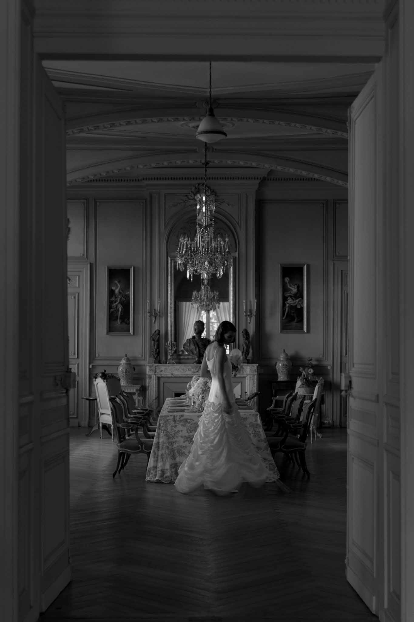 Bride from behind in ruffled gown inside grand chateau salon with chandelier and paneled walls in black and white