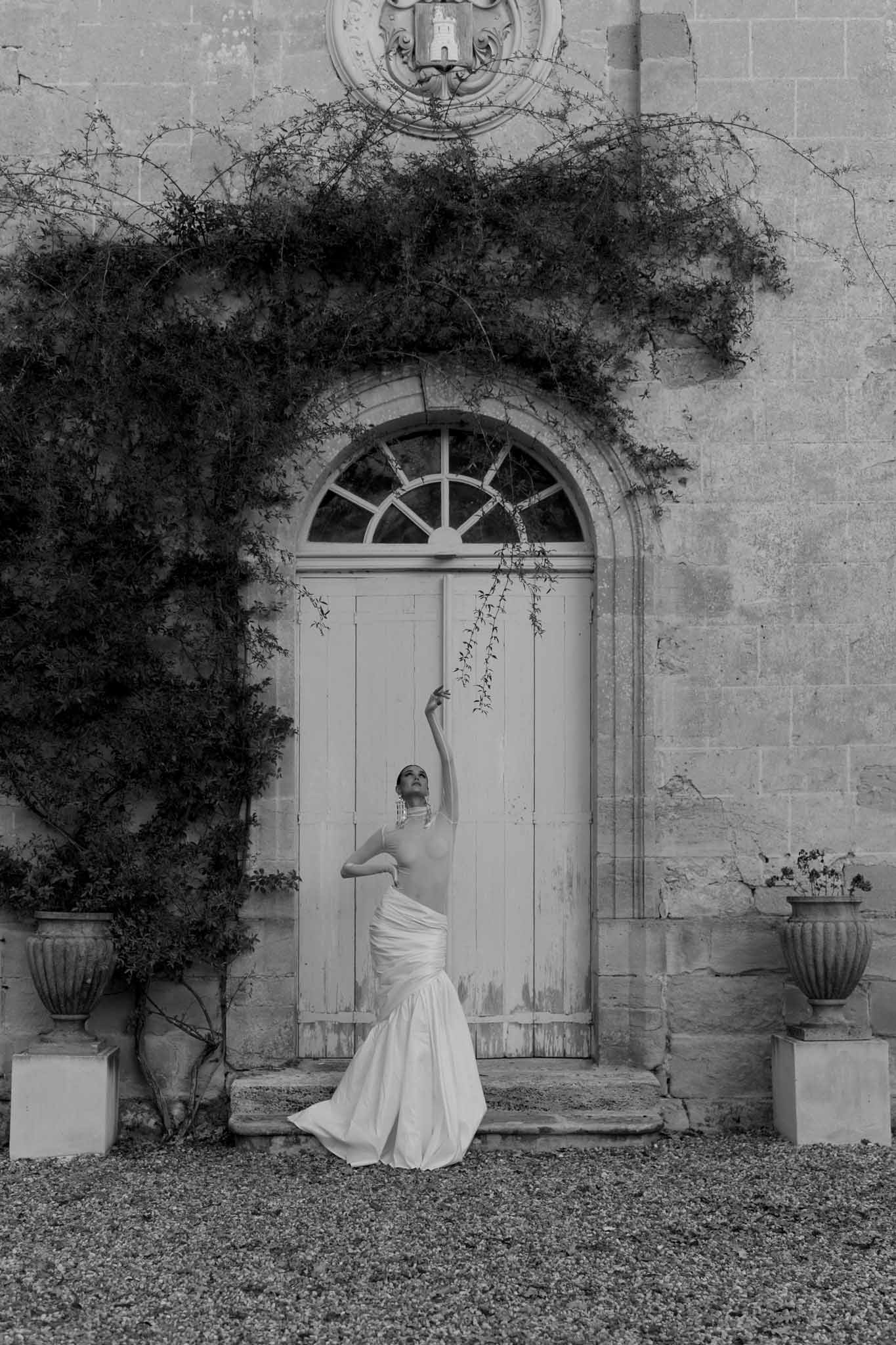 Black-and-white bridal portrait of bride posing dramatically before arched chateau doorway with trailing vines