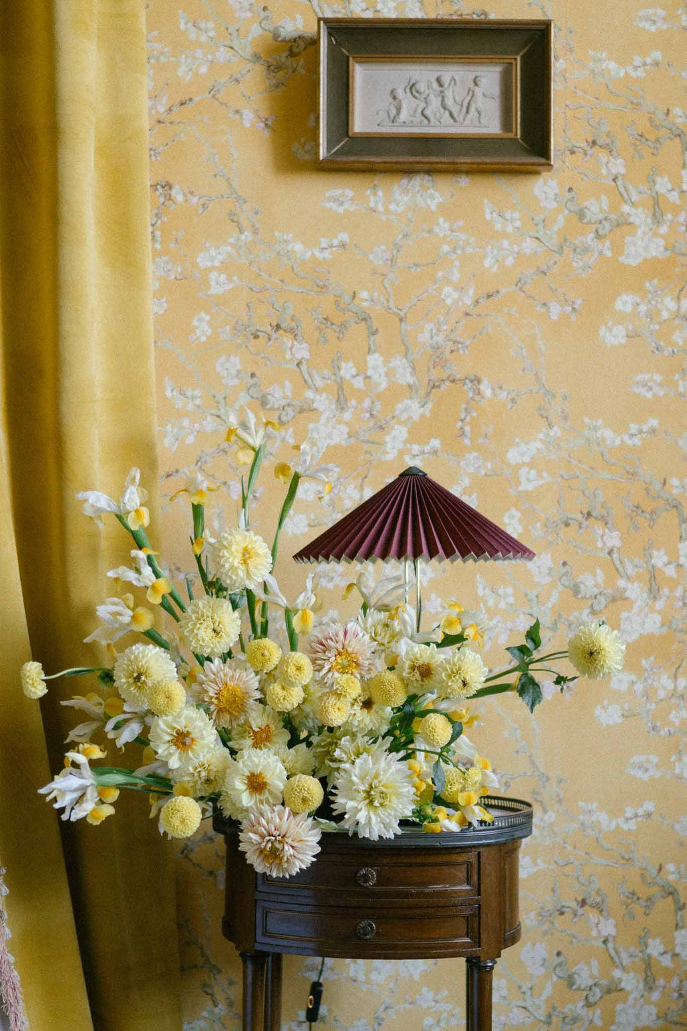 Overflowing yellow dahlia and iris arrangement on walnut table against ochre blossom wallpaper