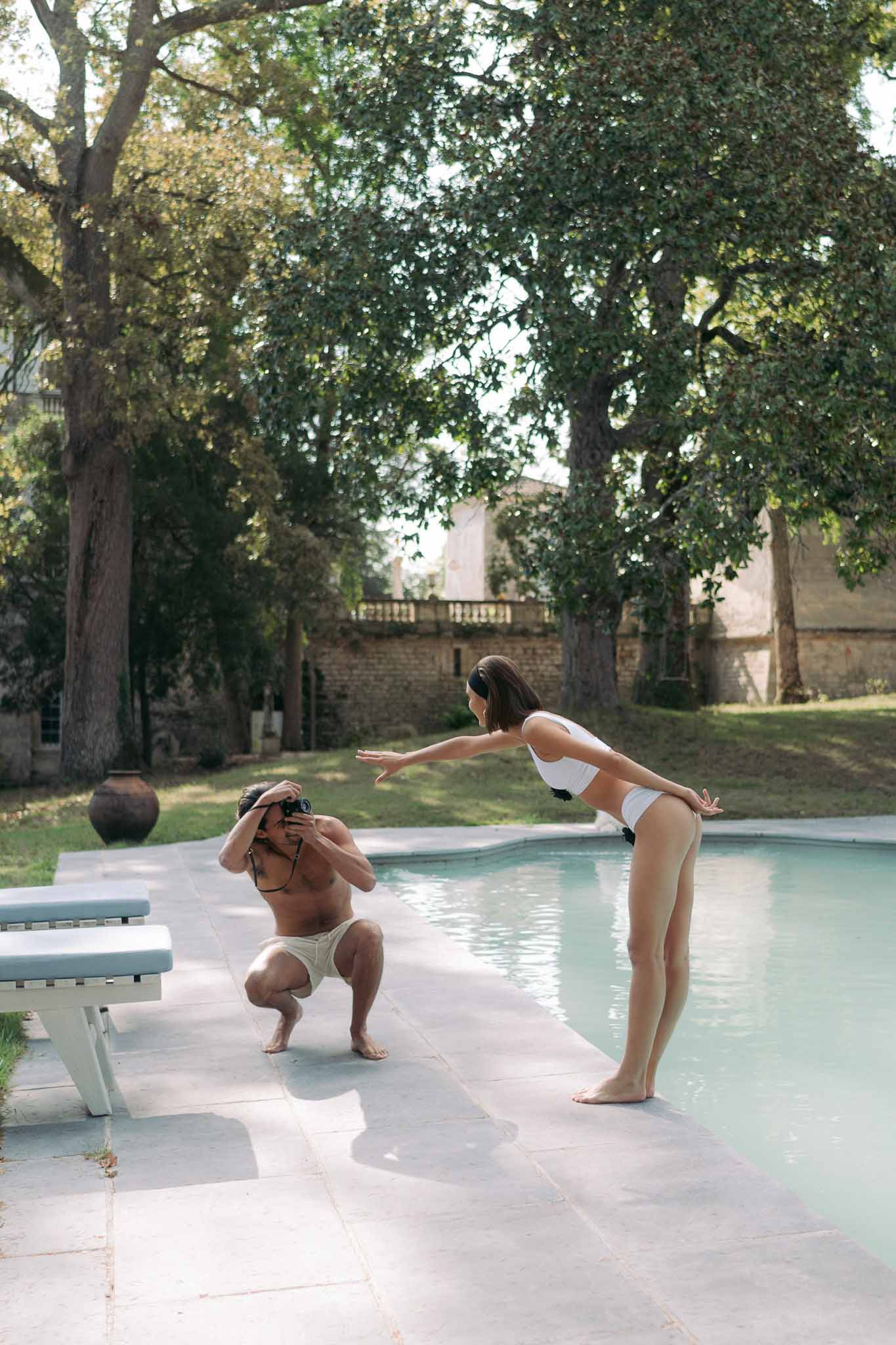 Couple at poolside groom crouching with film camera photographing bride in white bikini at estate pool