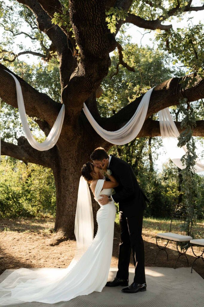 manoir de letang myrto events bride holding face groom hugging kissing ceremony altar under big tree