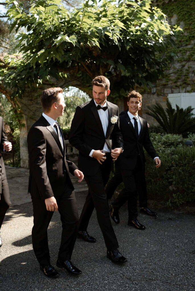 manoir de letang myrto events groom groomsmen walking holding tuxedo chatting garden