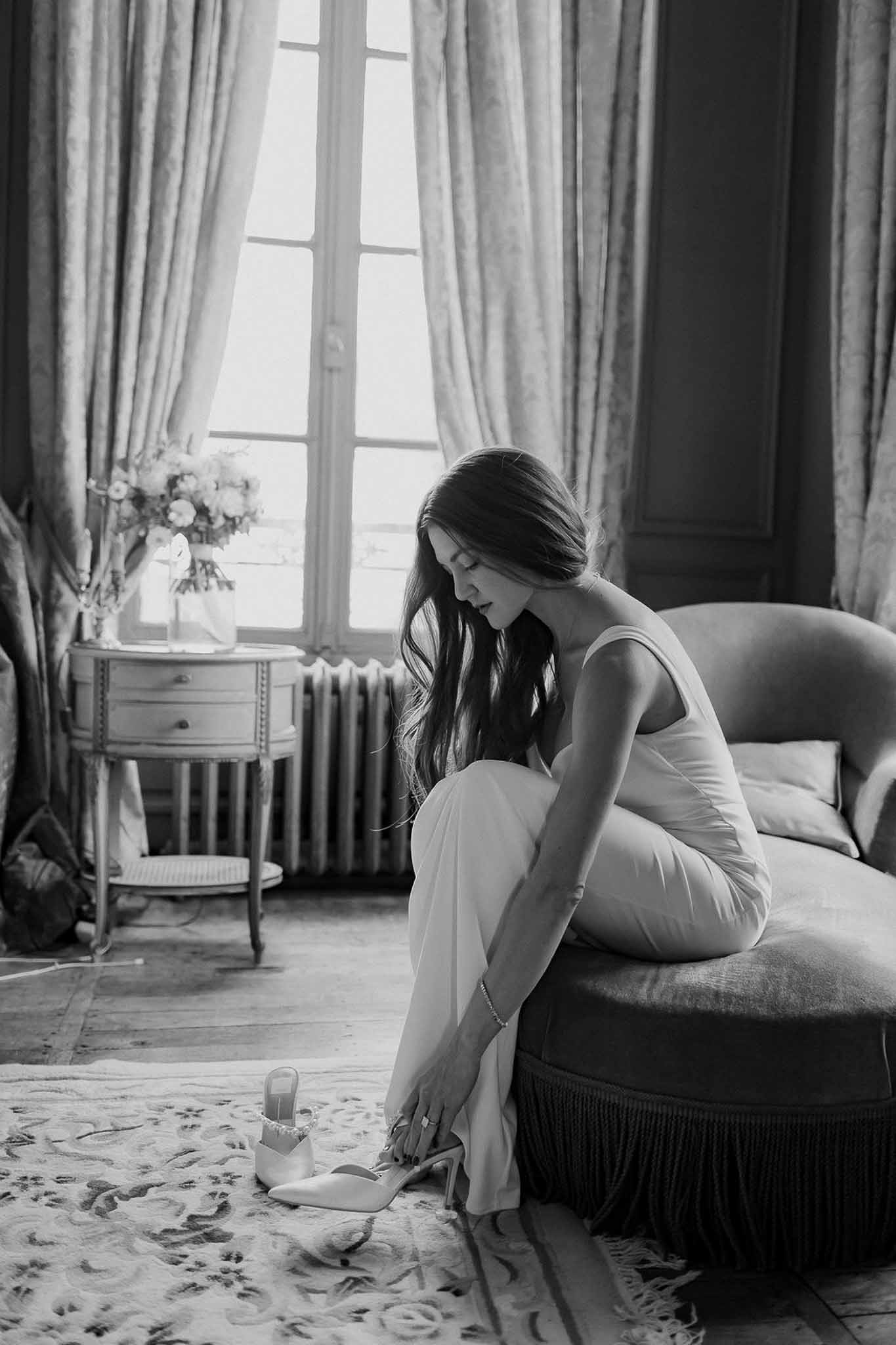 Black and white photo of bride seated on chaise longue putting on pointed-toe bridal heels in chateau room