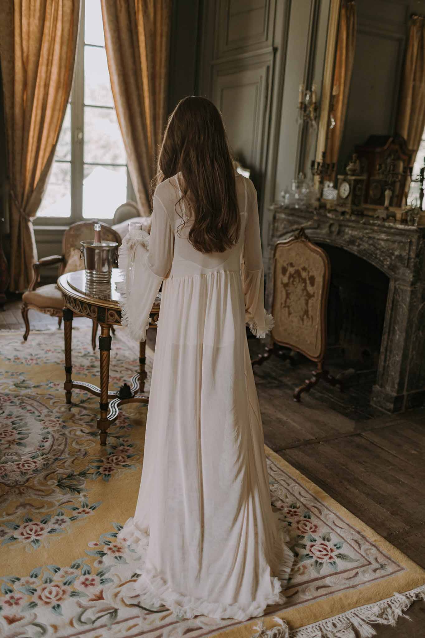 Bride in ivory robe with bell sleeves holding champagne flute in sage-panelled chateau salon