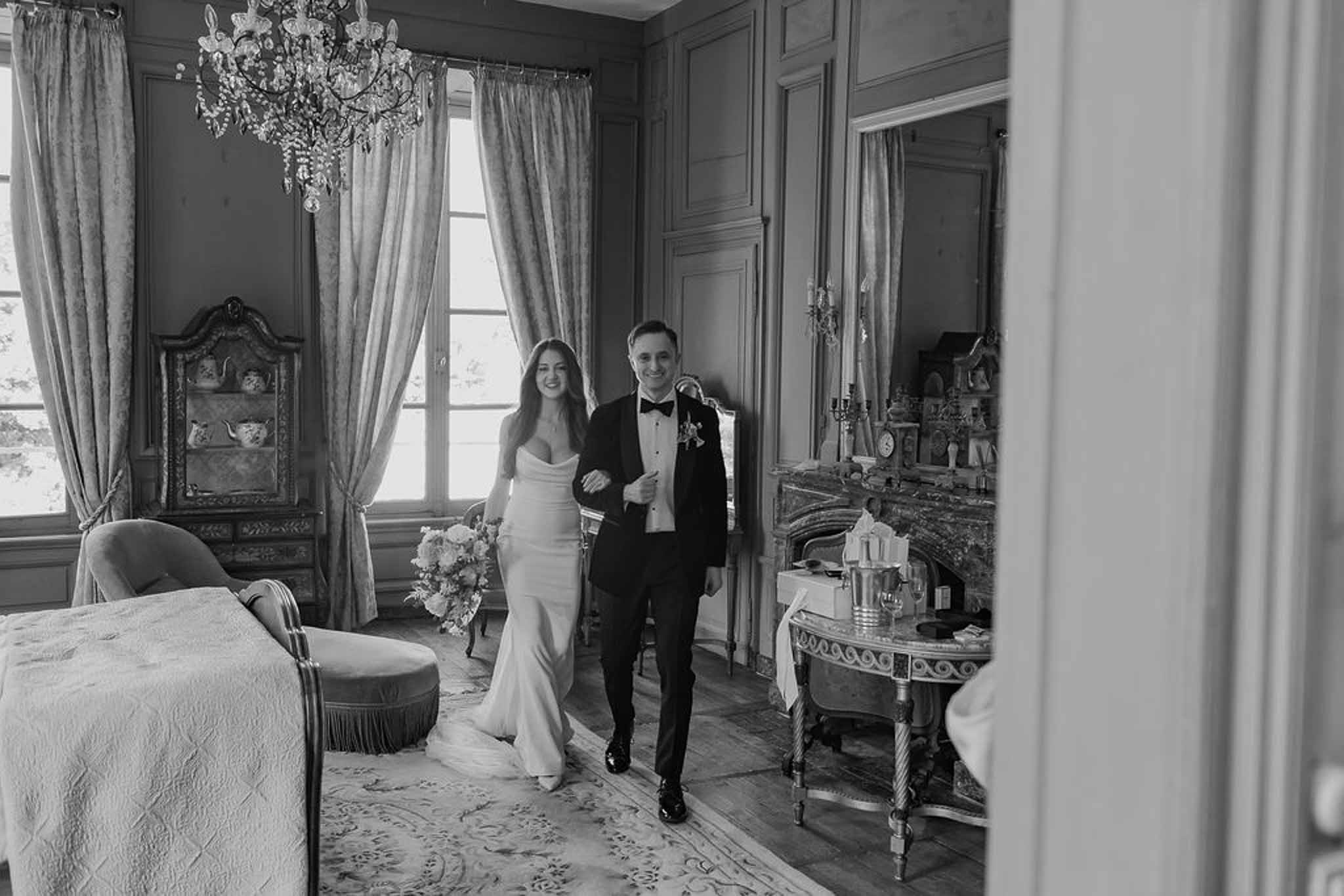 Black and white portrait of couple walking through chateau room with chandelier and drapes