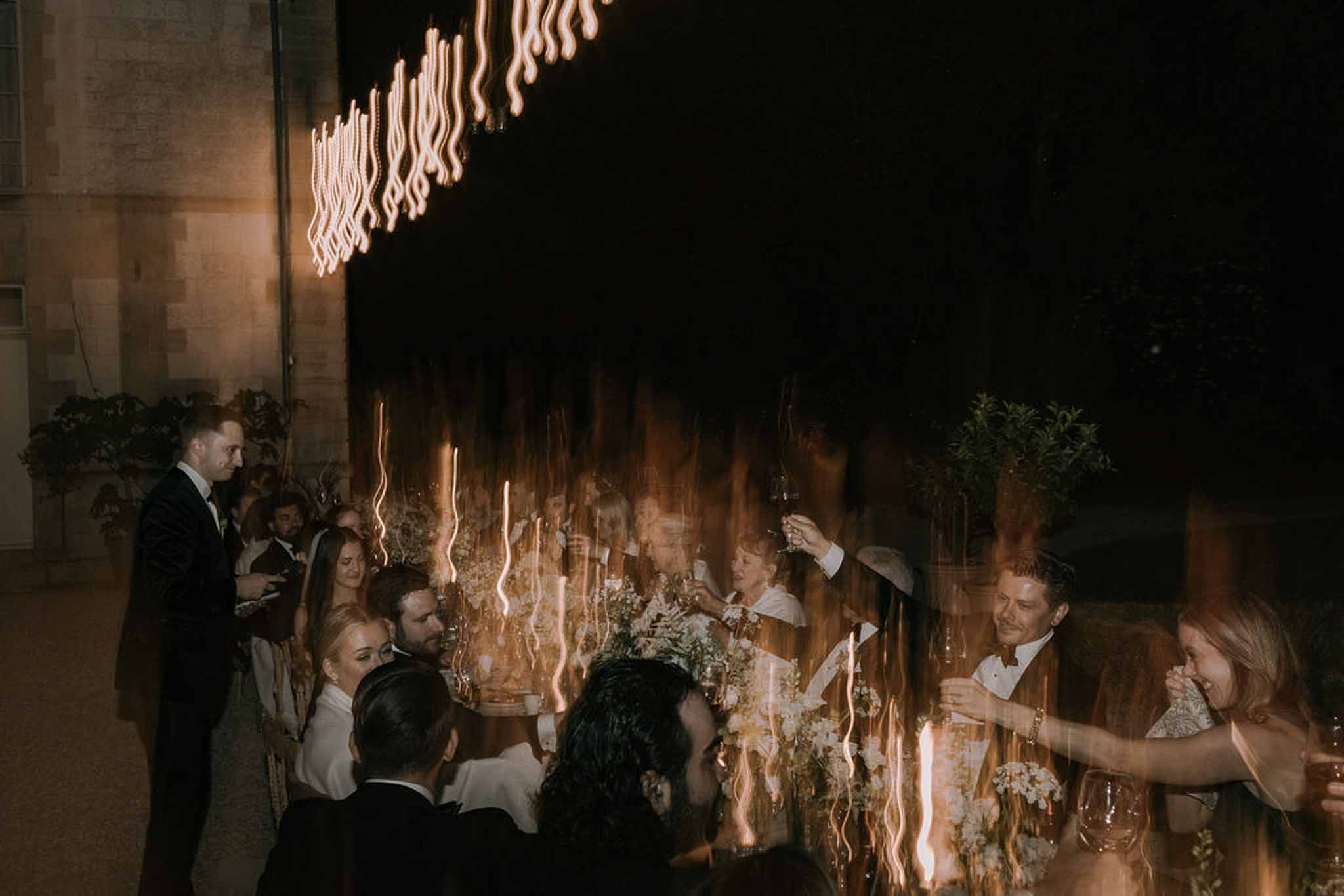 Long-exposure evening reception with guests raising champagne glasses under streaked warm pendant lights in a courtyard