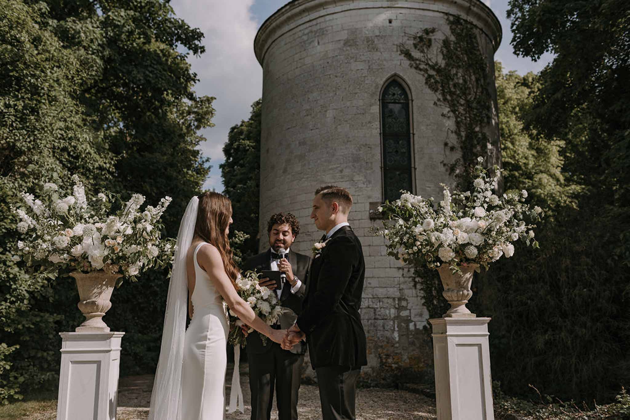 Bride and groom hold hands at altar before stone tower flanked by white rose and hydrangea arrangements