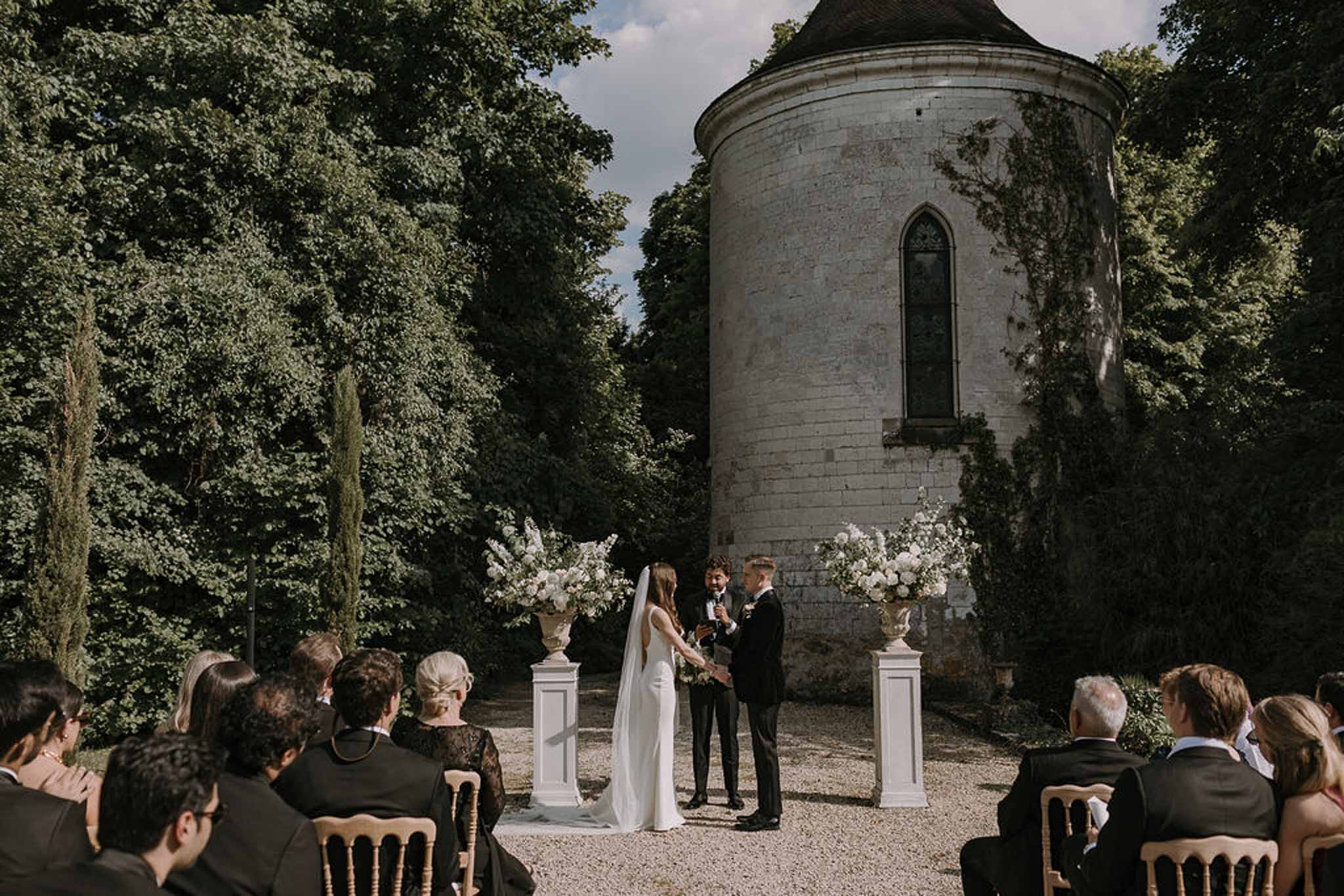 Outdoor wedding ceremony in front of a round stone chateau tower with white floral urns and guests in gold chairs