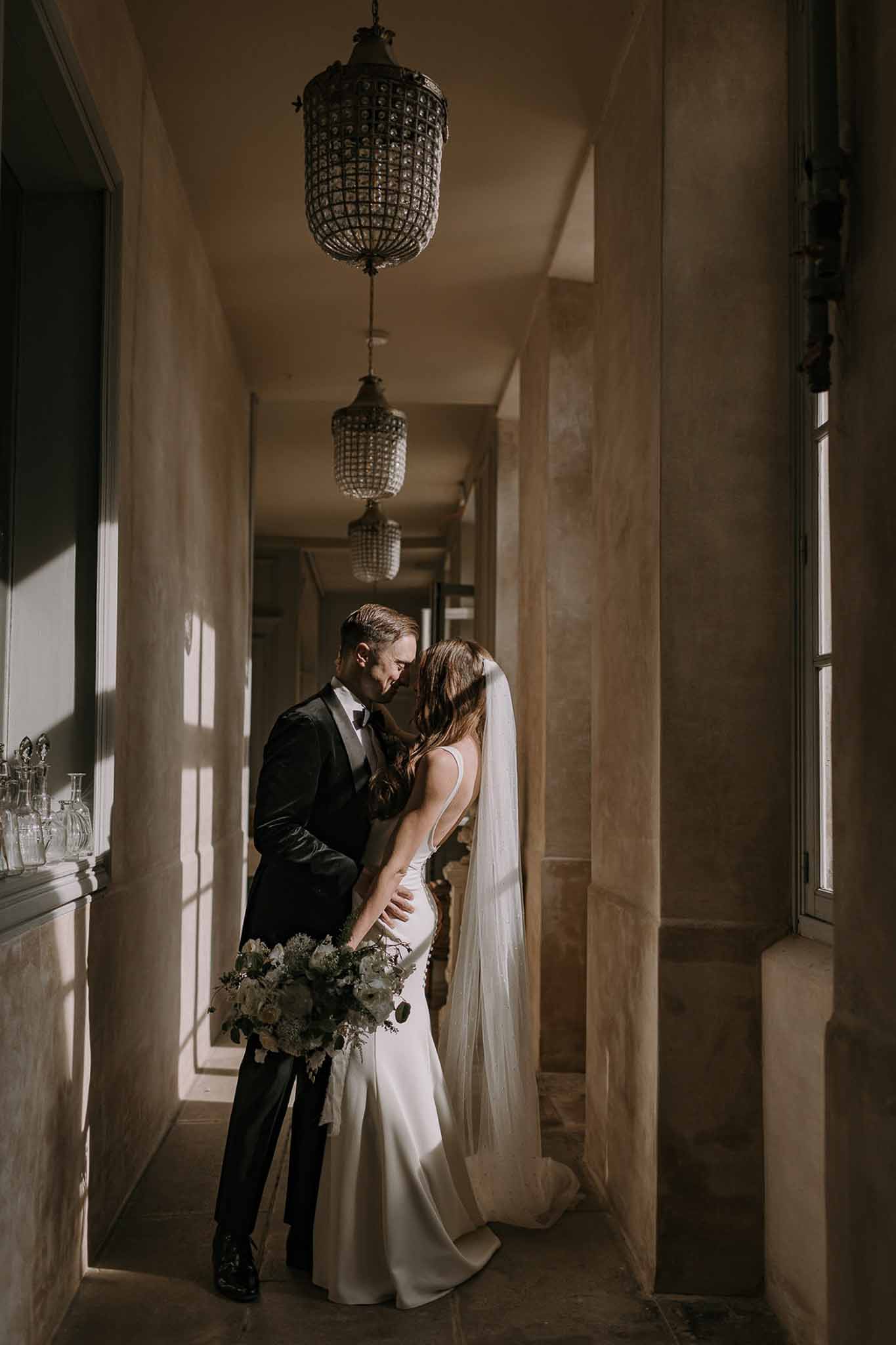 Couple touching foreheads in chandelier-lit chateau corridor, bride with ivory veil and cream bouquet