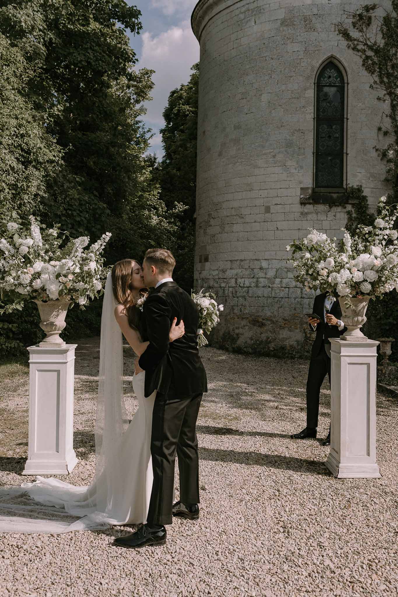 Bride and groom share first kiss at outdoor chateau ceremony flanked by white floral urns